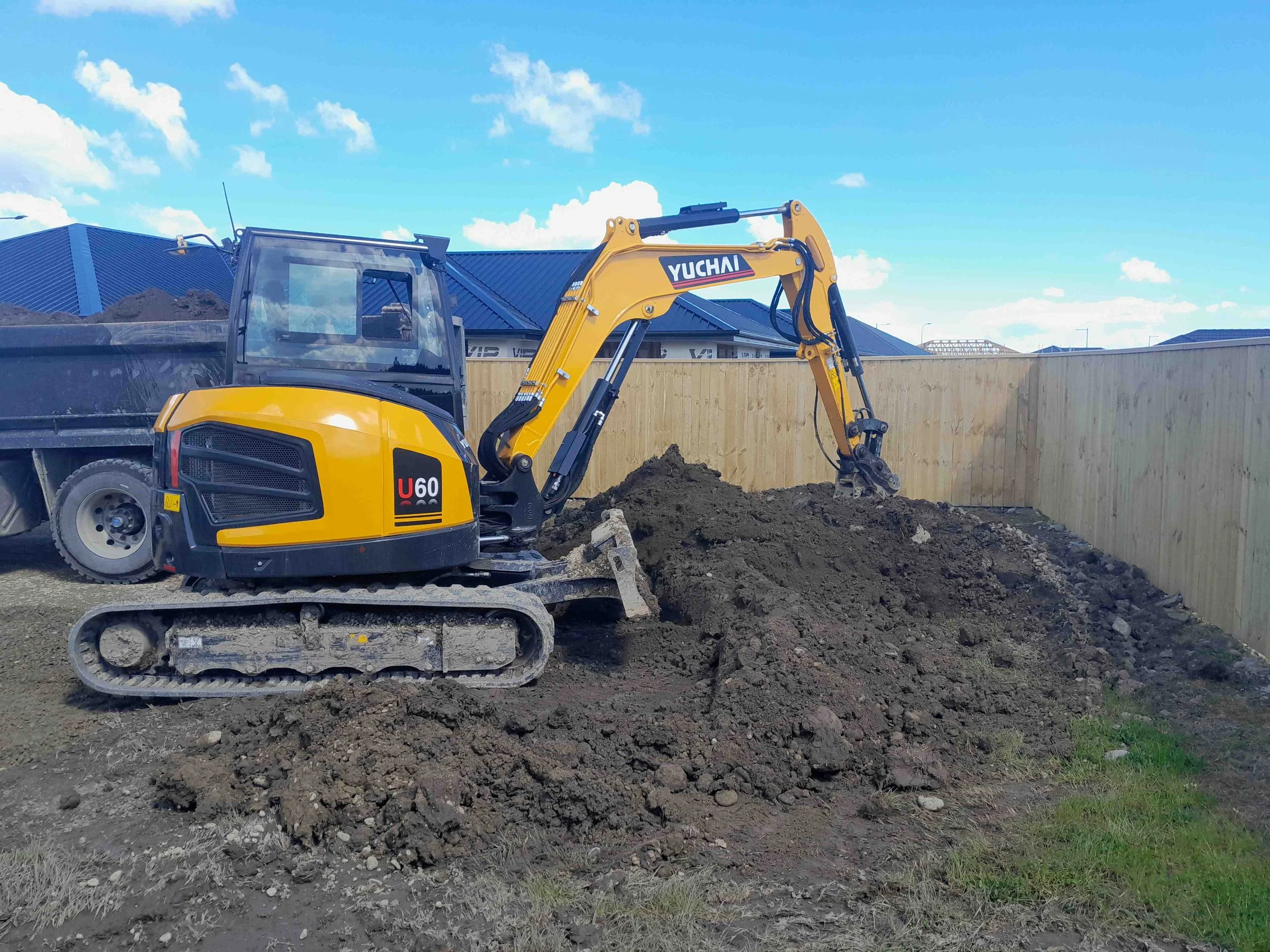 A yellow Yuchai mini excavator digger on a construction site with dark soil pile in front, a wooden fence, and a blue sky with some scattered clouds in the background.