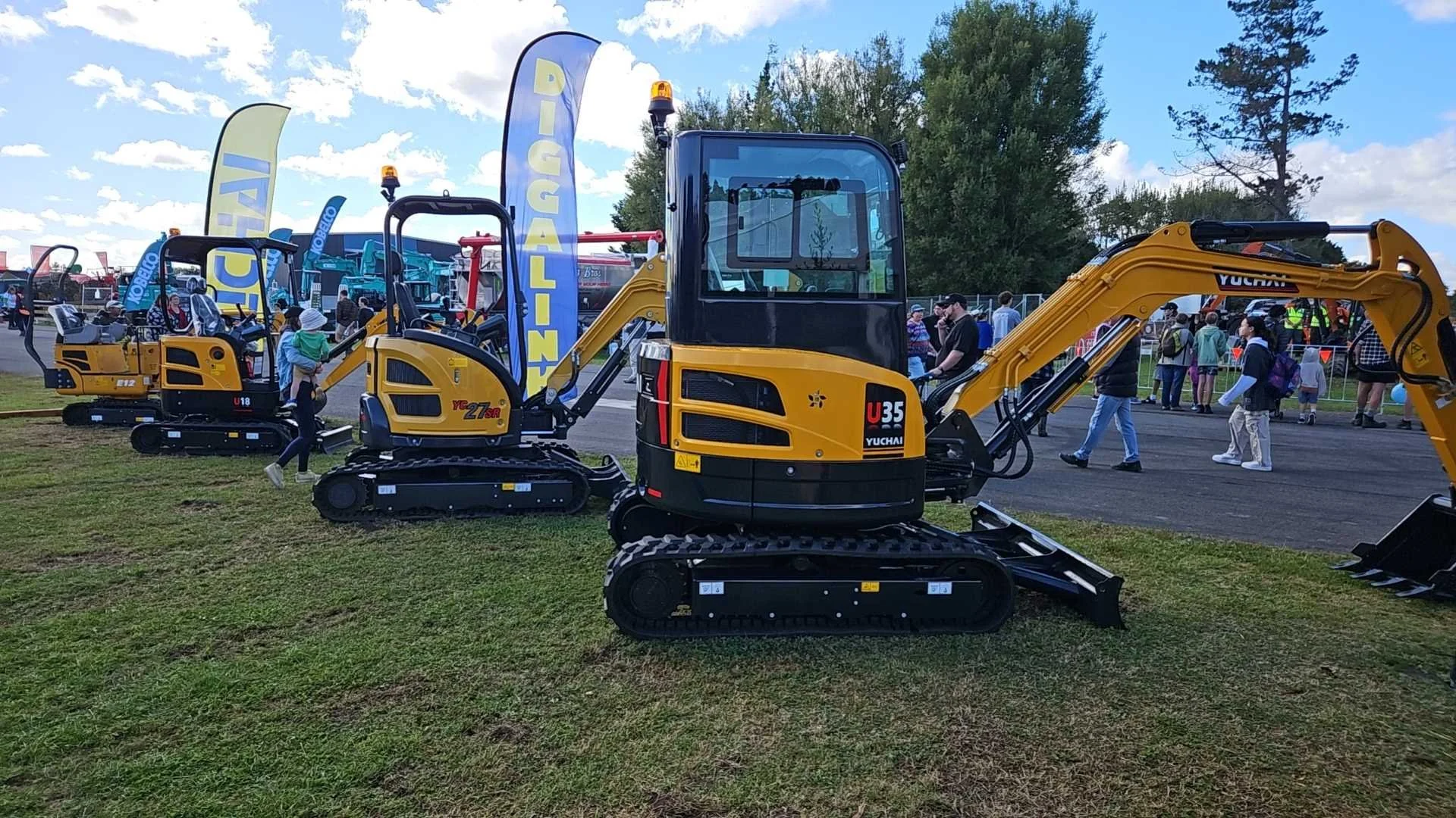 At an outdoor event, multiple yellow and black mini excavators are displayed on a grassy area with a crowd of people walking around and looking at them. There are flags and banners advertising the excavator brands, and trees can be seen in the backgr