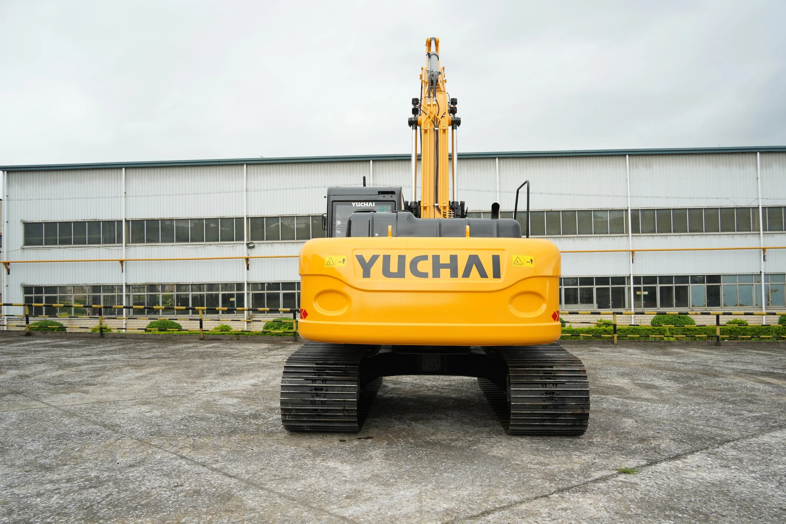 Yellow Yuchai excavator parked outdoors in front of large industrial building with glass windows.