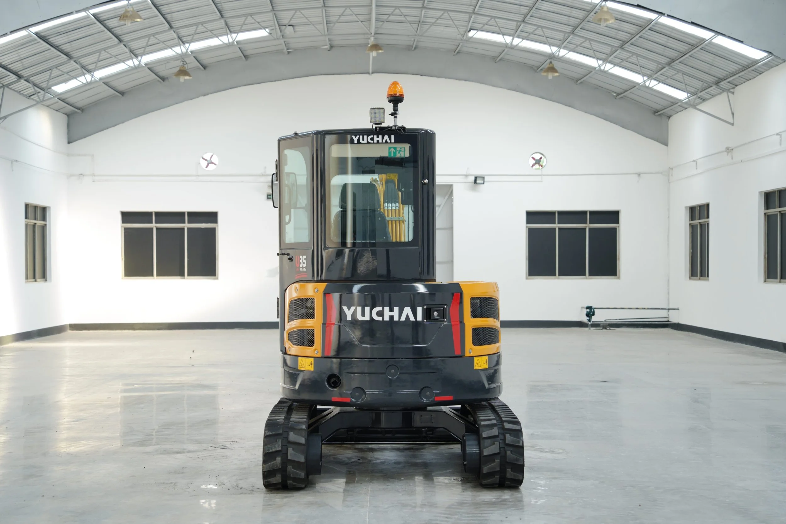 A Yuchai compact excavator with tracks is positioned in an indoor space with white walls, windows, a gray floor, and a curved metal ceiling. The front of the excavator is facing the camera.