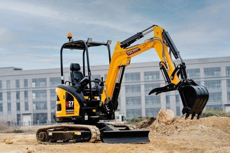Small yellow and black excavator working on a construction site with a modern building in the background