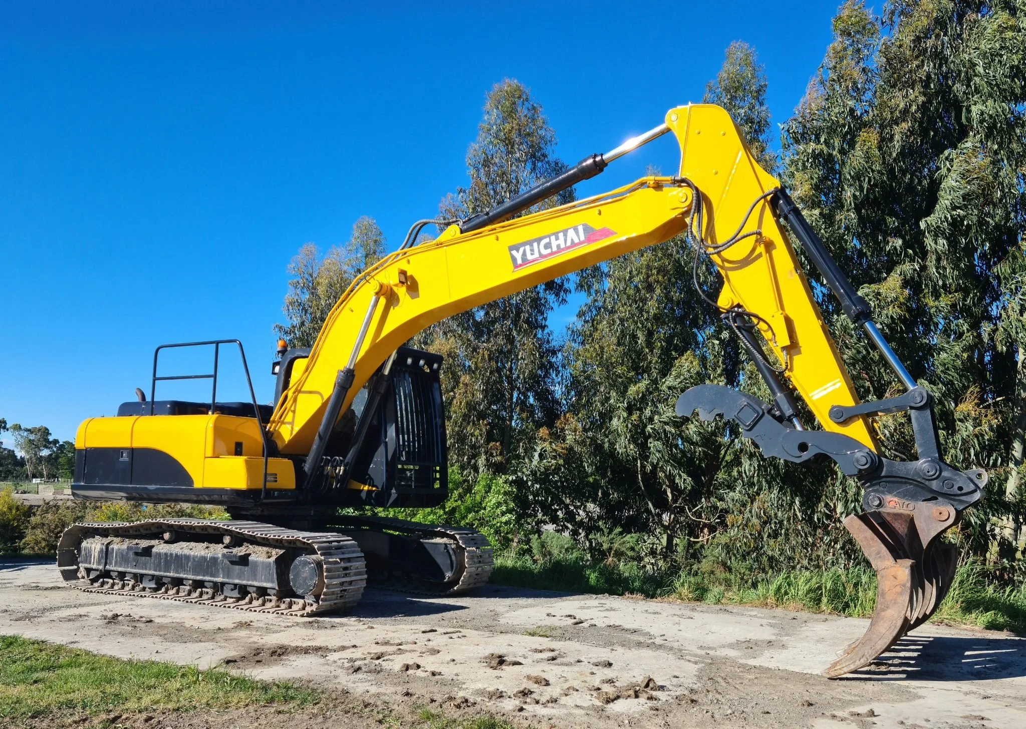 A yellow Yuchai excavator with black tracked wheels and hydraulic arm, parked on dirt ground with a backdrop of green trees and a clear blue sky.
