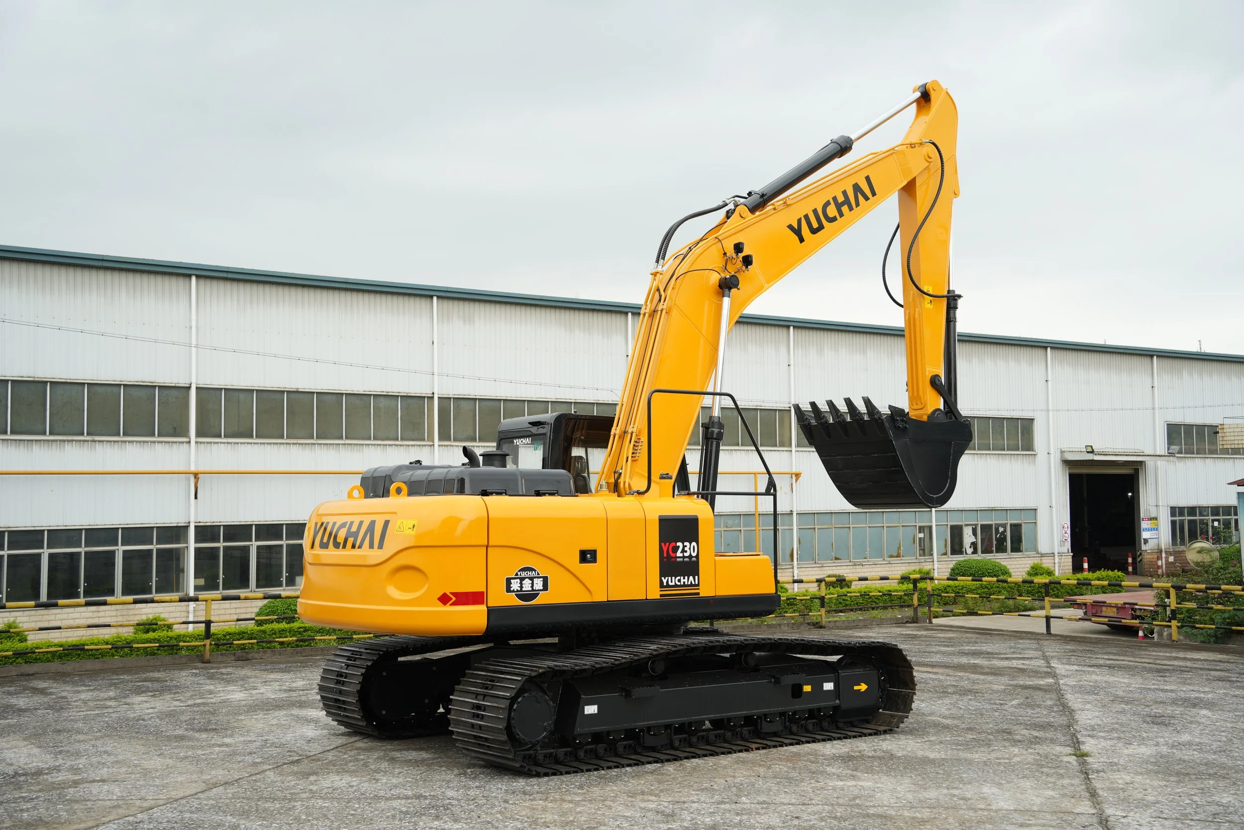 Yellow excavator with black caterpillar tracks parked outside a large industrial building, under an overcast sky.