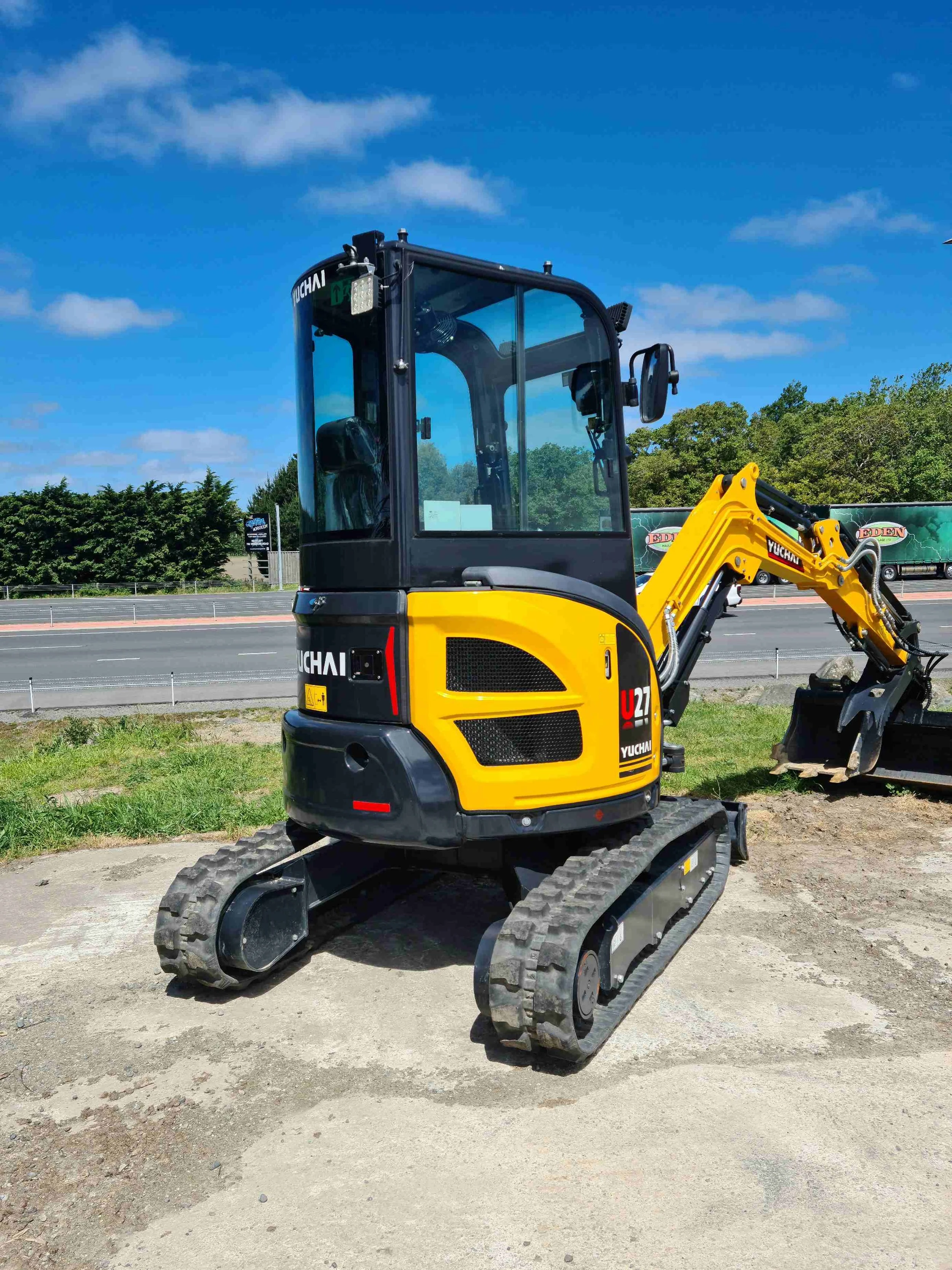 A small yellow and black tracked excavator with the brand name Yuchai on the side, parked outdoors on a patch of ground with grass and dirt, under a blue sky with some clouds.