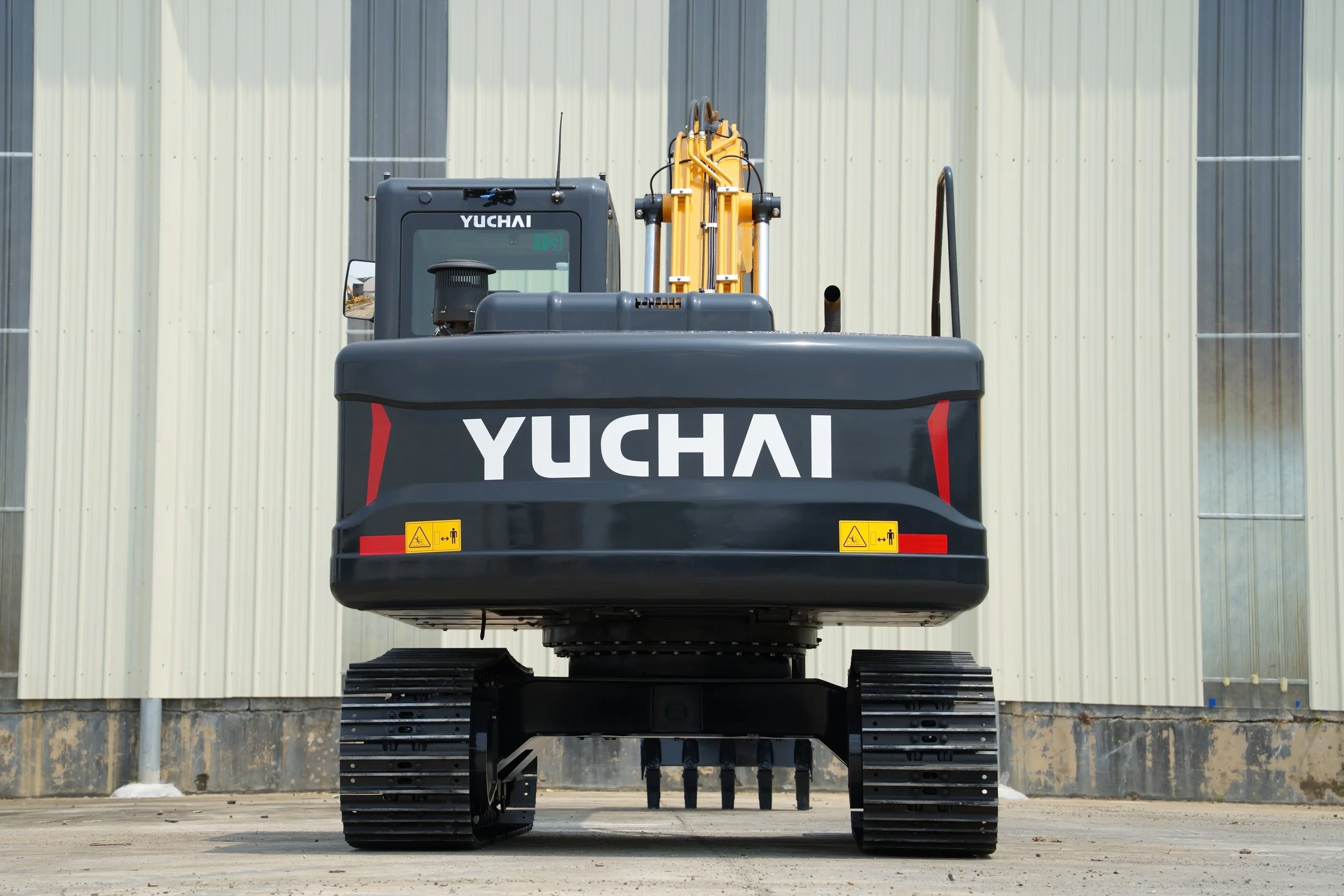 Front view of a large Yuchai excavator with black and yellow parts, parked outside a building with vertical paneled metal siding.