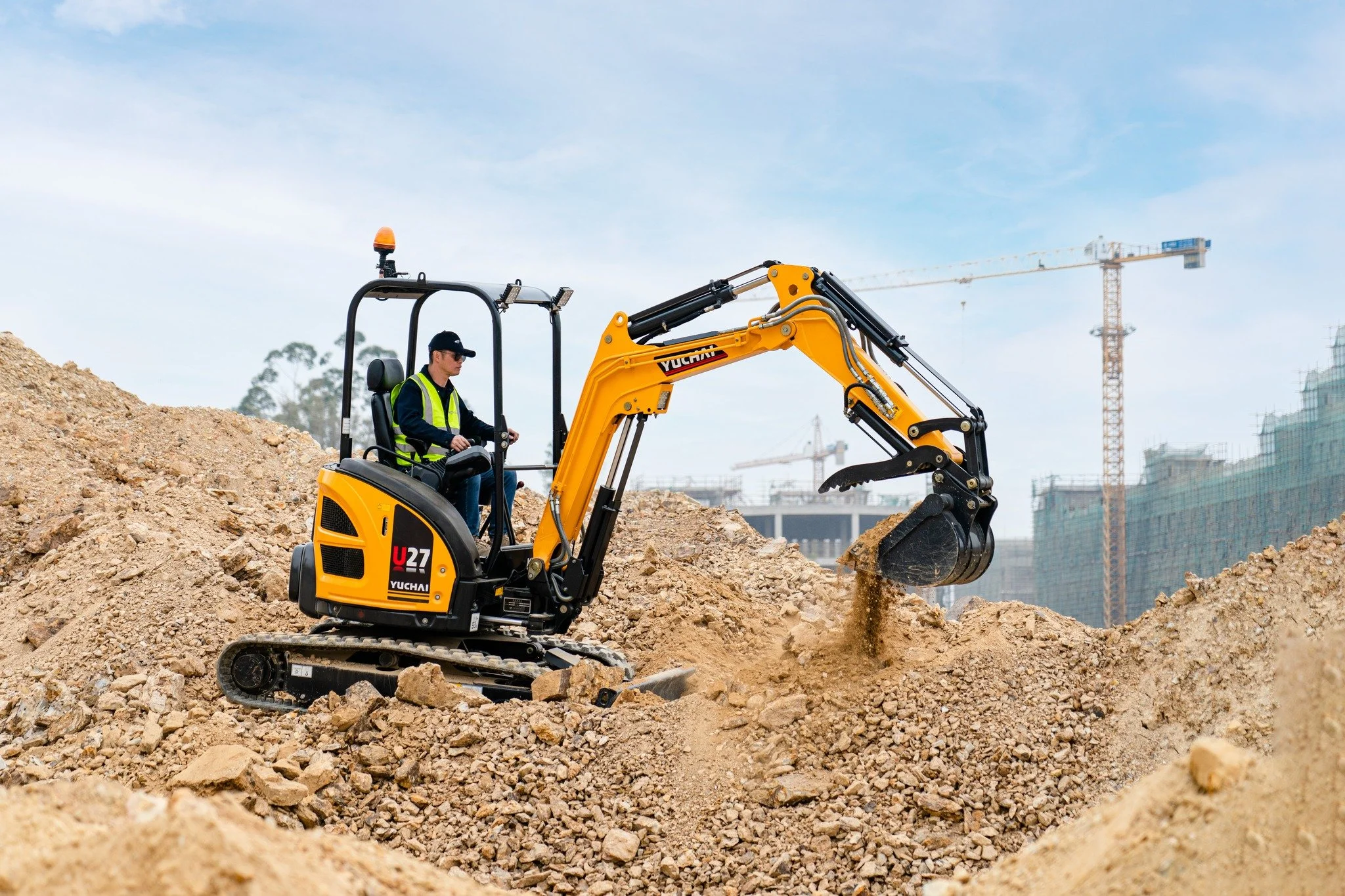A construction worker operating a yellow mini excavator on a dirt site with construction cranes in the background.