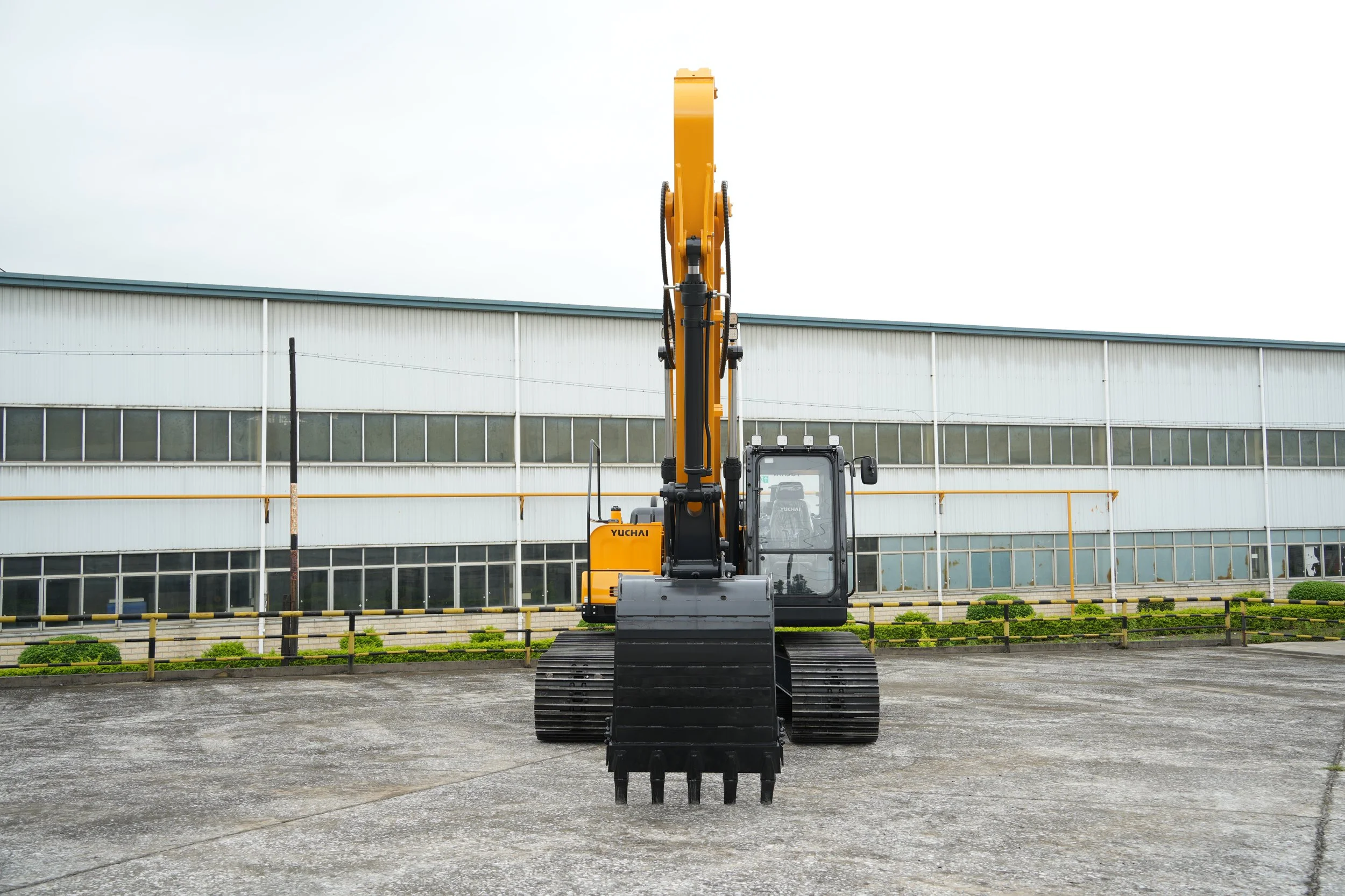 A yellow excavator sits on a gravel lot in front of a large industrial building with horizontal siding, and a decorative fence surrounds the area.
