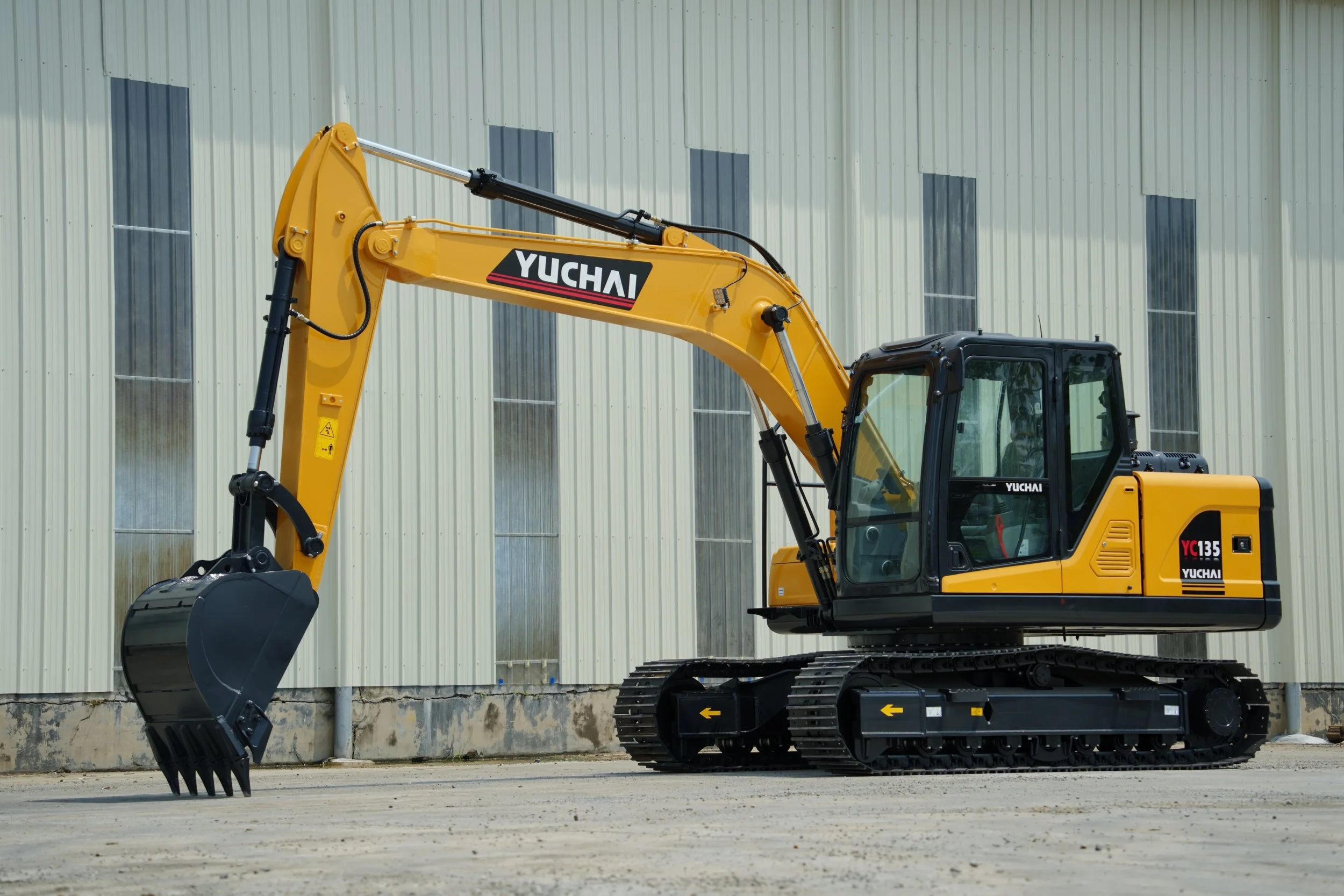 A yellow Yuchai excavator with a black bucket attachment parked outside a large building with metal siding and windows.