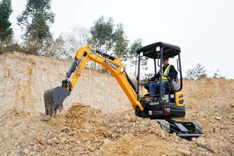 A man operating a yellow mini excavator on a construction site with dirt and rocks, surrounded by trees.