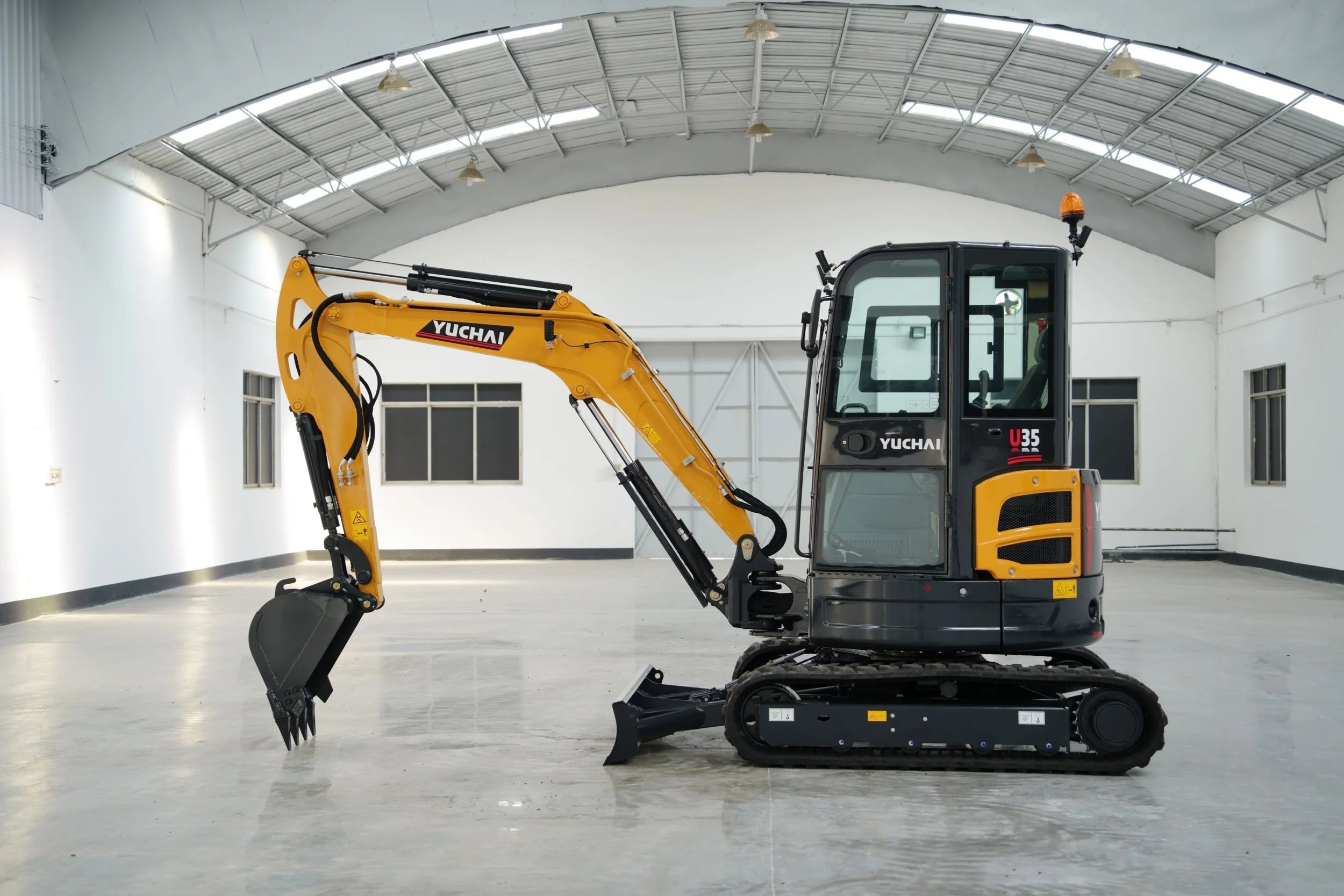 Small yellow and black Yuchai excavator inside a spacious, well-lit warehouse with a curved metal ceiling and concrete floor.