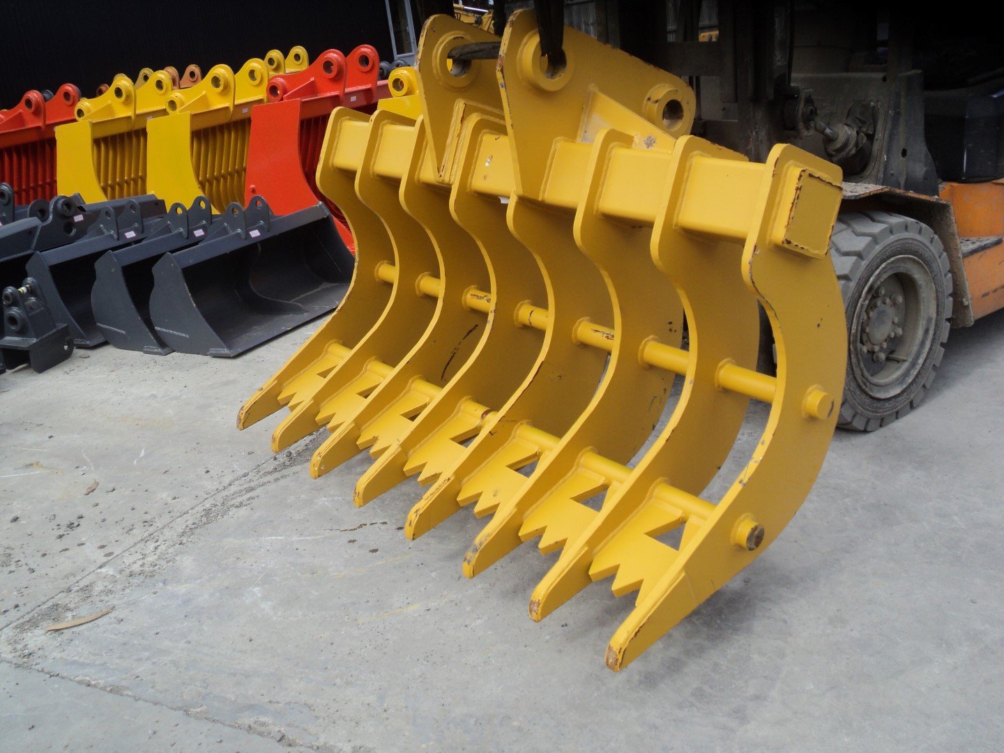 Multiple large excavator buckets in yellow, black, and red colors, lined up on concrete ground, with a forklift near a construction equipment warehouse in the background.