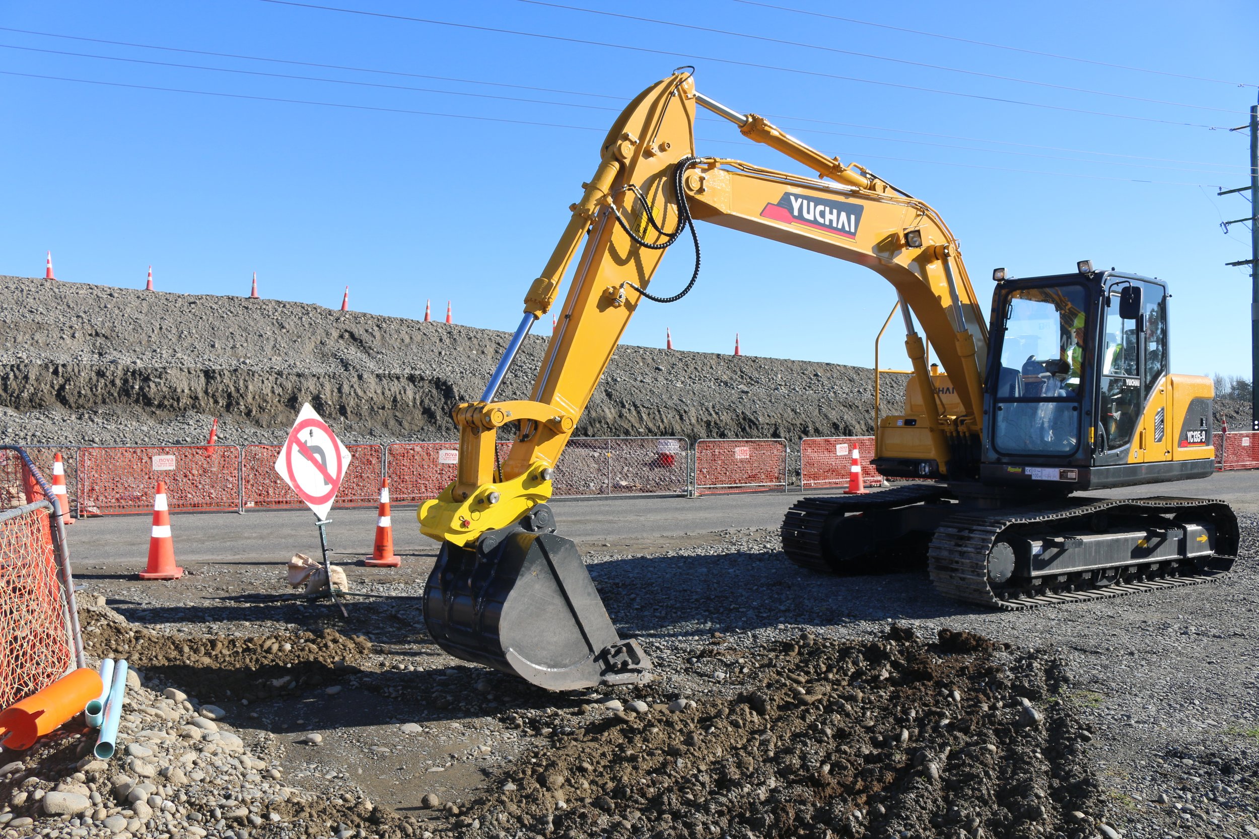 A yellow Yuchai excavator at a construction site, digging into dirt with orange and white safety cones and fencing around. The sky is clear and blue, with power lines overhead.
