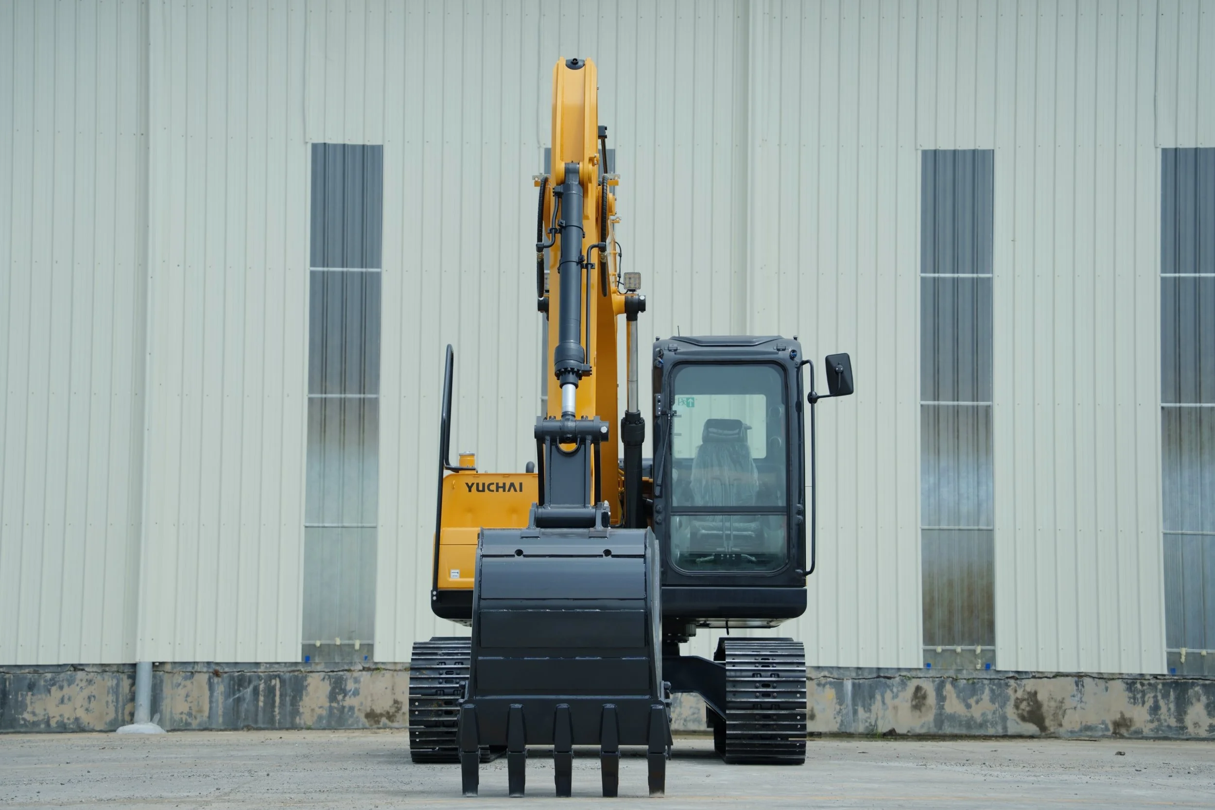A yellow Yuchai excavator parked in front of a light-colored metal building with vertical panels.
