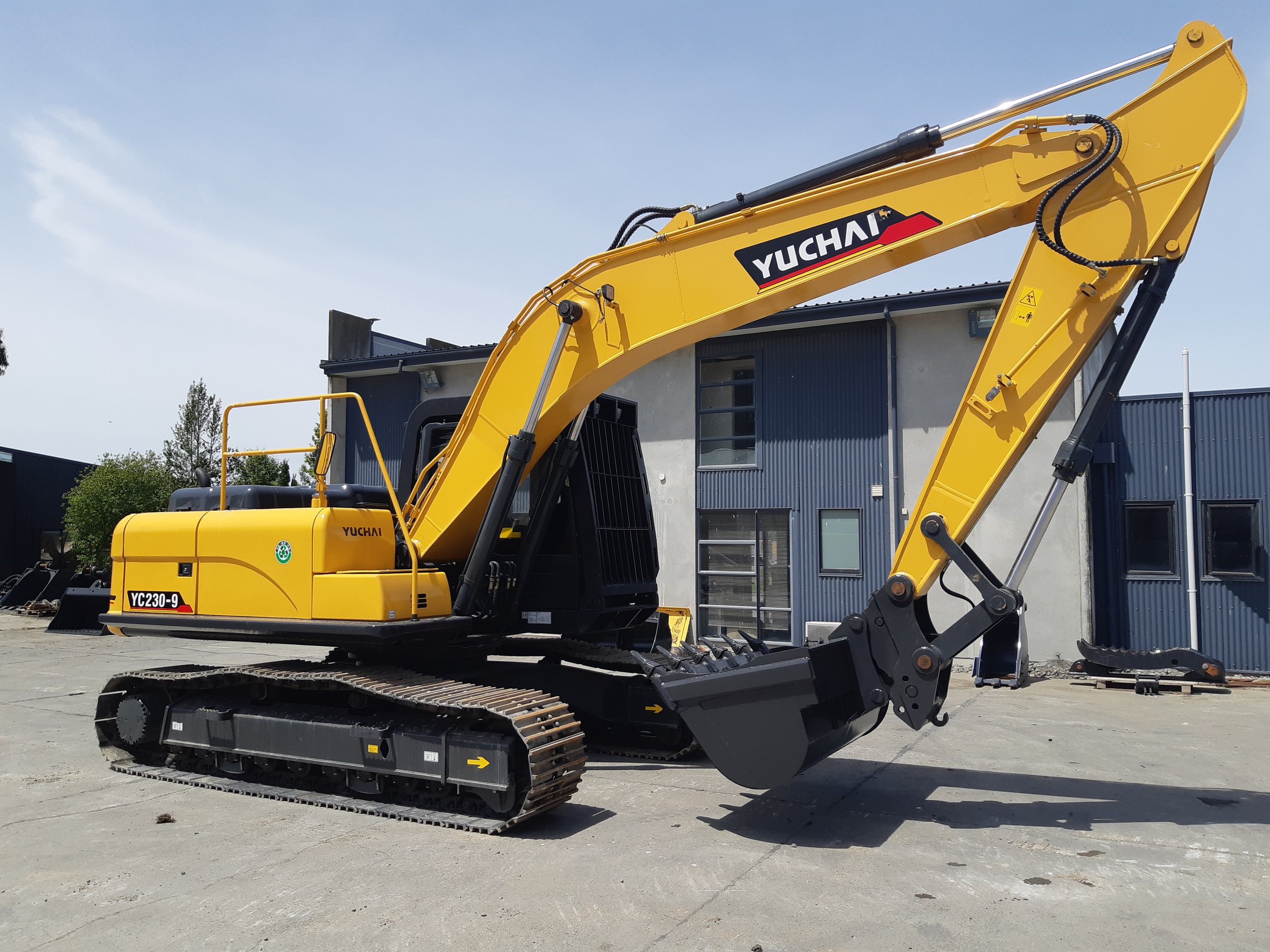 Yellow Yuchai excavator with track system parked outdoors with building in background.
