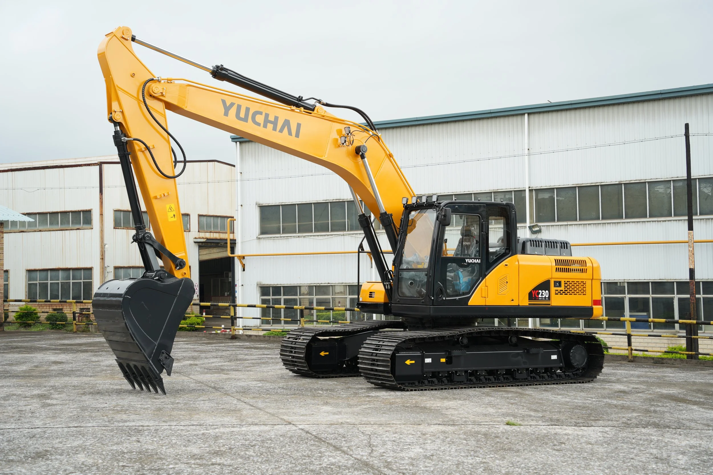 A yellow Yuchai excavator with black tracks parked on a concrete surface in front of industrial buildings.