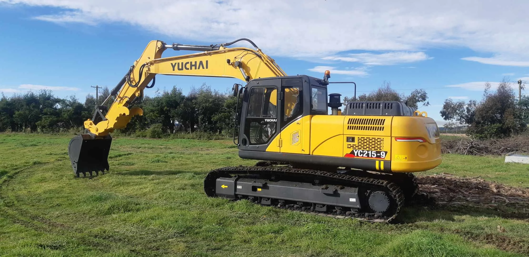 A yellow Yuchai YC215-9 excavator operating on a grassy field with trees and power lines in the background under a partly cloudy sky.