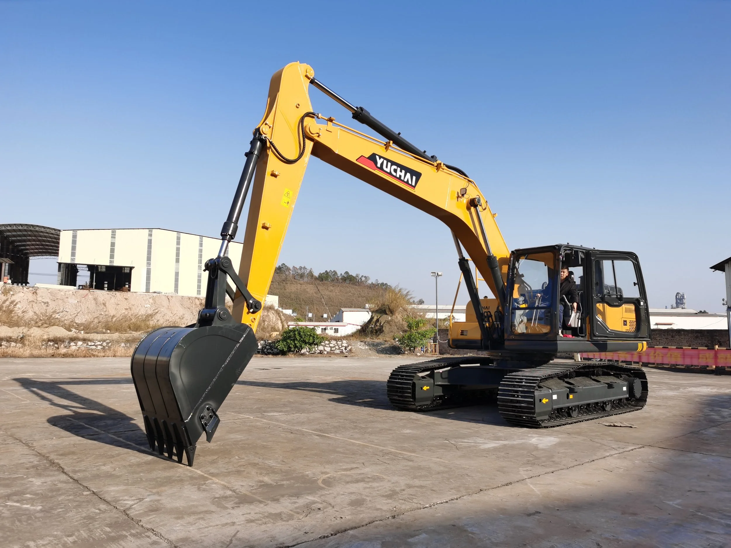 A large yellow construction excavator with black tracks and a black cabin, with a person inside, parked on a concrete surface under a clear blue sky.
