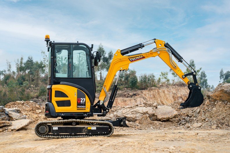 Small yellow and black excavator working on dirt and rocks at a construction site with trees in the background.