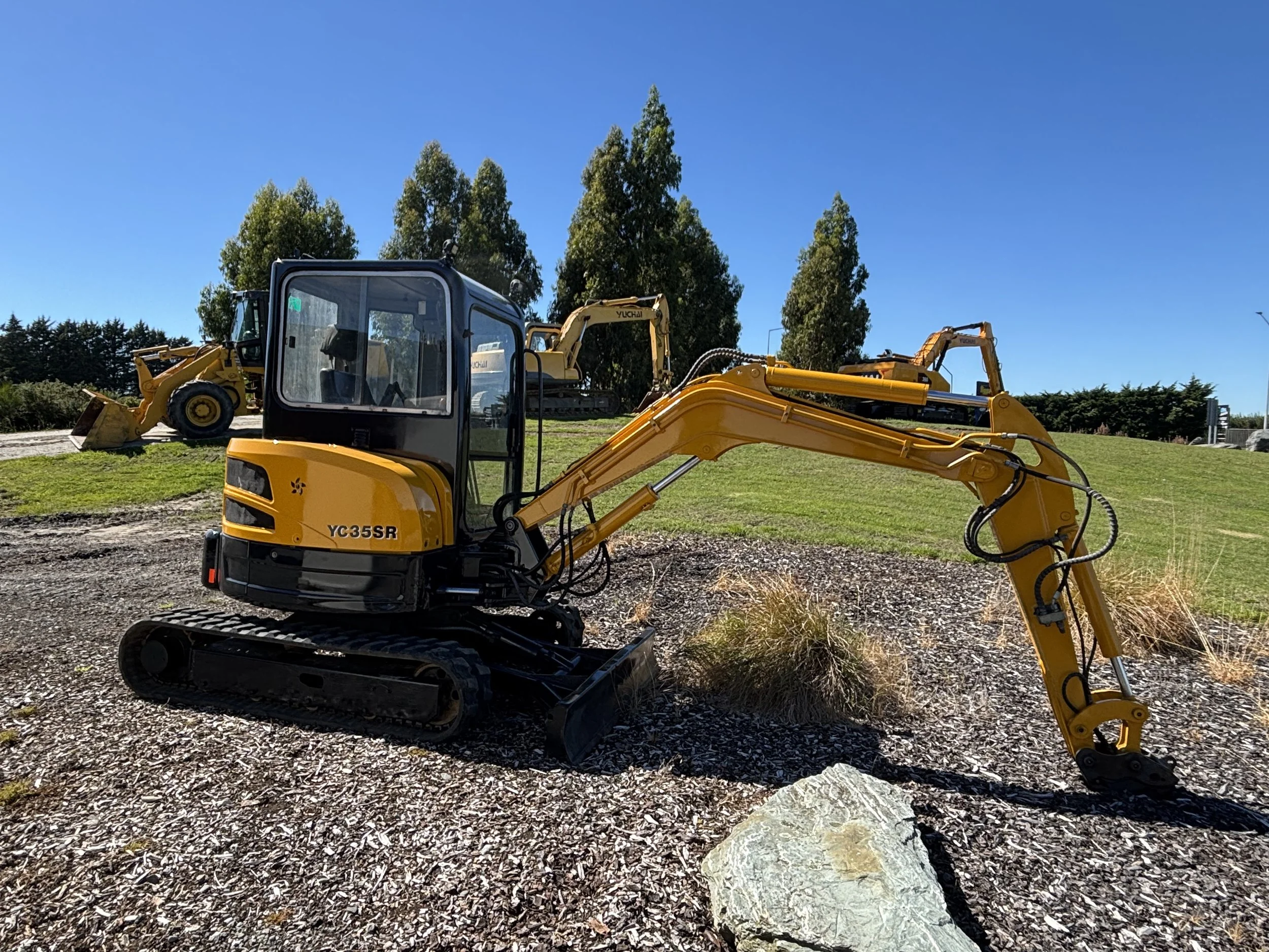 Yellow mini excavator on gravel, with a larger bulldozer in the background, and tall trees under a clear blue sky.