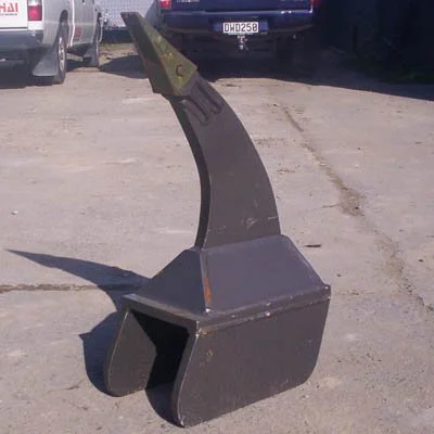 A large black excavator bucket with a curved shank and a pointed attachment on top, placed on a gravel lot with parked cars in the background.