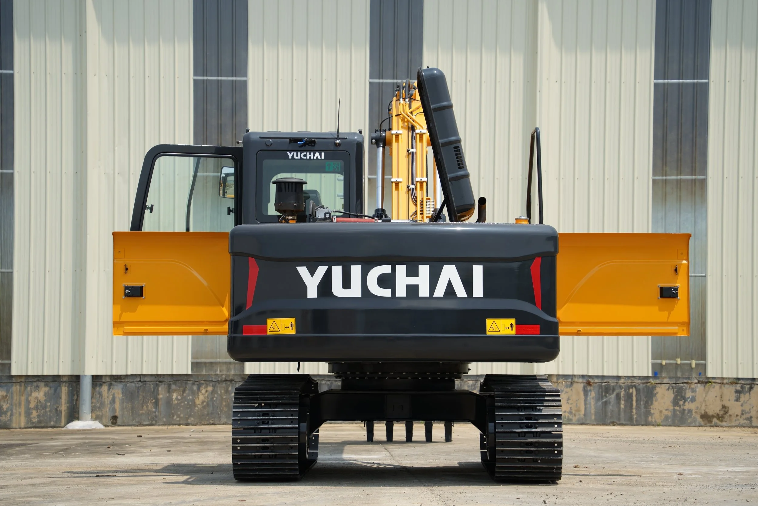 A yellow and black Yuchai mini excavator with black rubber tracks parked outside on a concrete surface, against a metallic wall with vertical panels.