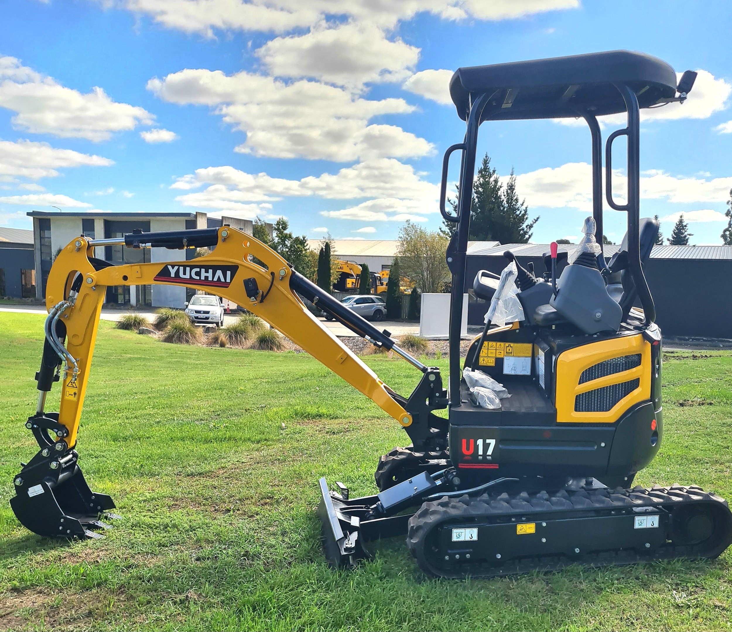 A yellow and black Yuchai compact excavator with a digging arm on a grassy area, with buildings, parked cars, and trees in the background under a blue sky with scattered clouds.