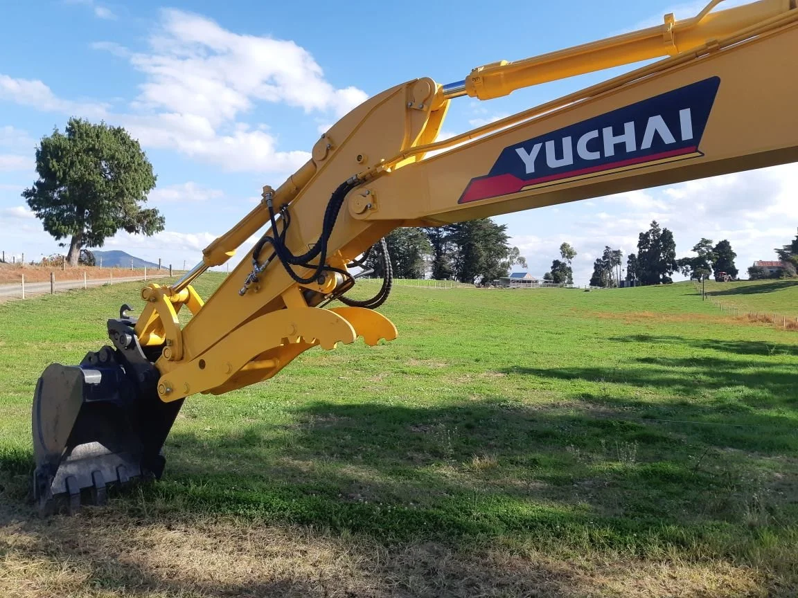 Yellow Yuchai excavator arm with bucket attachment on green grass field, blue sky with white clouds, trees, and distant buildings.