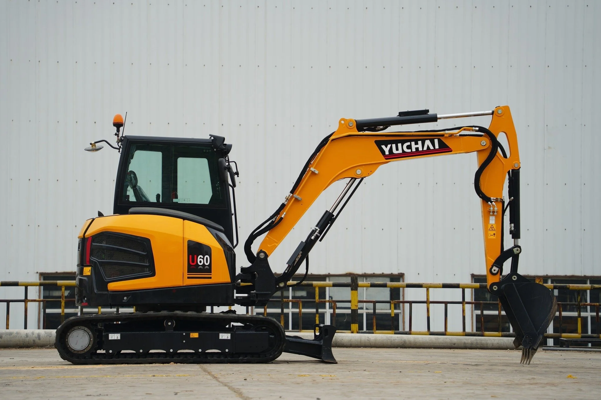 Yellow and black Yuchai mini excavator with tracked wheels and hydraulic arm, parked outdoors on concrete surface in front of a large white industrial building with small windows.