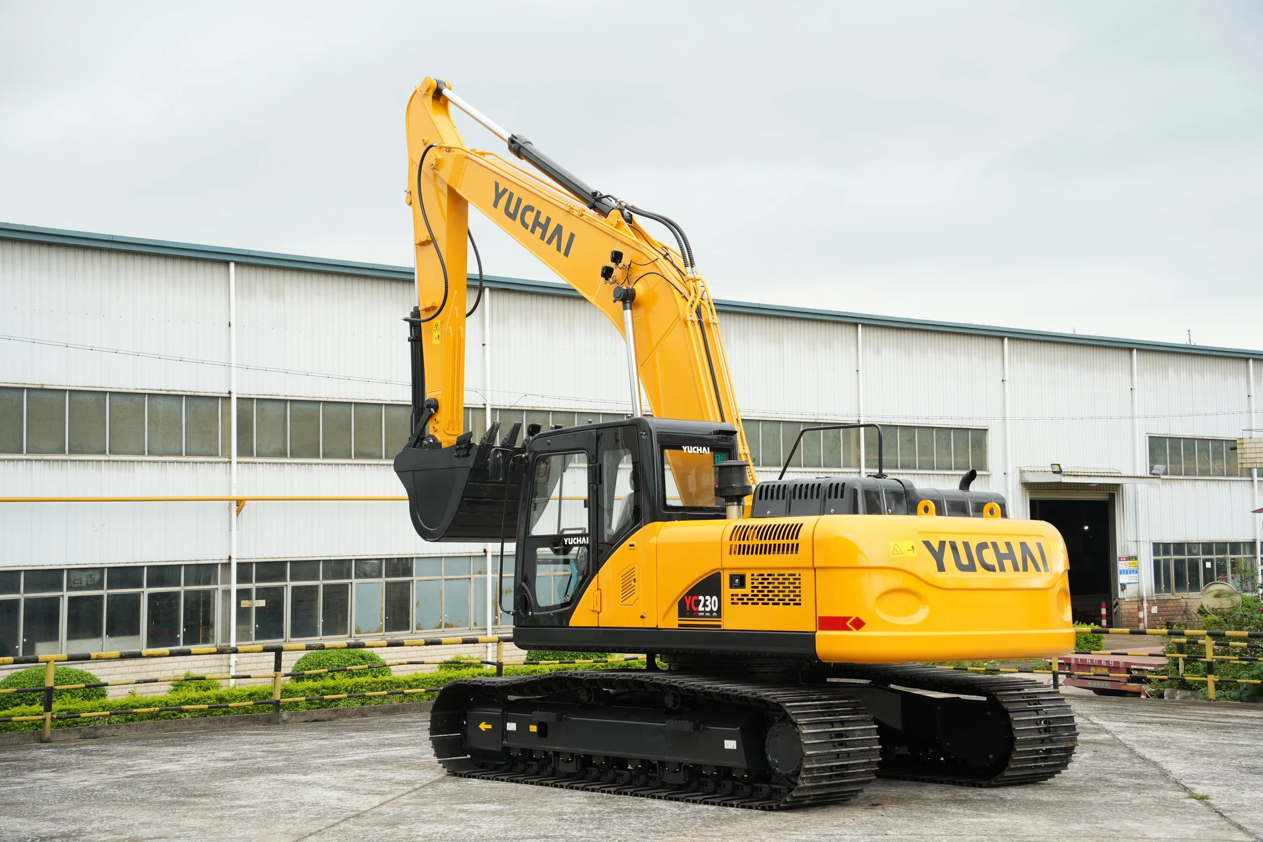 Yellow Yuchai excavator parked outdoors in front of a large industrial building with glass windows, under an overcast sky.