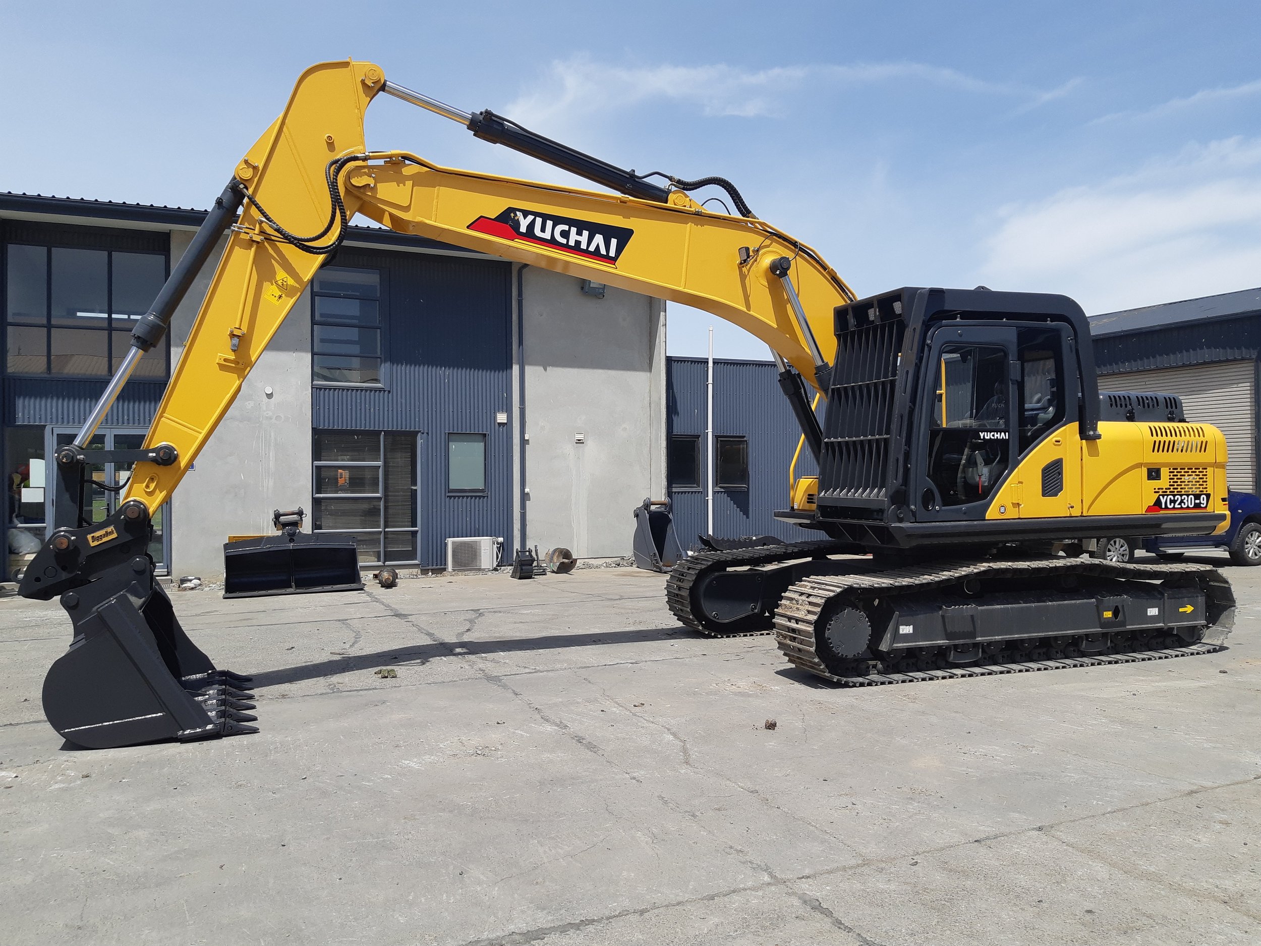 A yellow Yuchi excavator with a black bucket attachment is parked on a paved area outside a modern building with dark gray walls and large windows. The excavator's arm is extended, and the sky is partly cloudy.