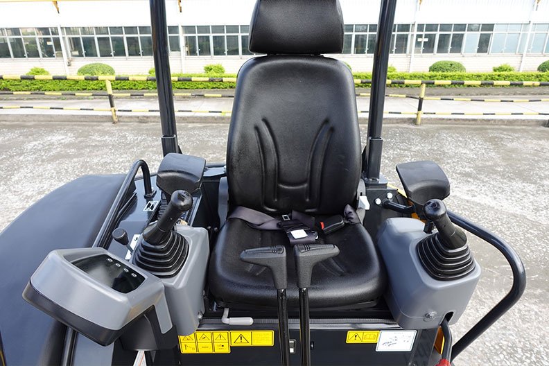 The image shows the operator's seat and control panel of a construction or farm vehicle, likely a skid steer or compact loader, with two joystick controls on either side of the chair, outside a building with large windows and a parking lot.