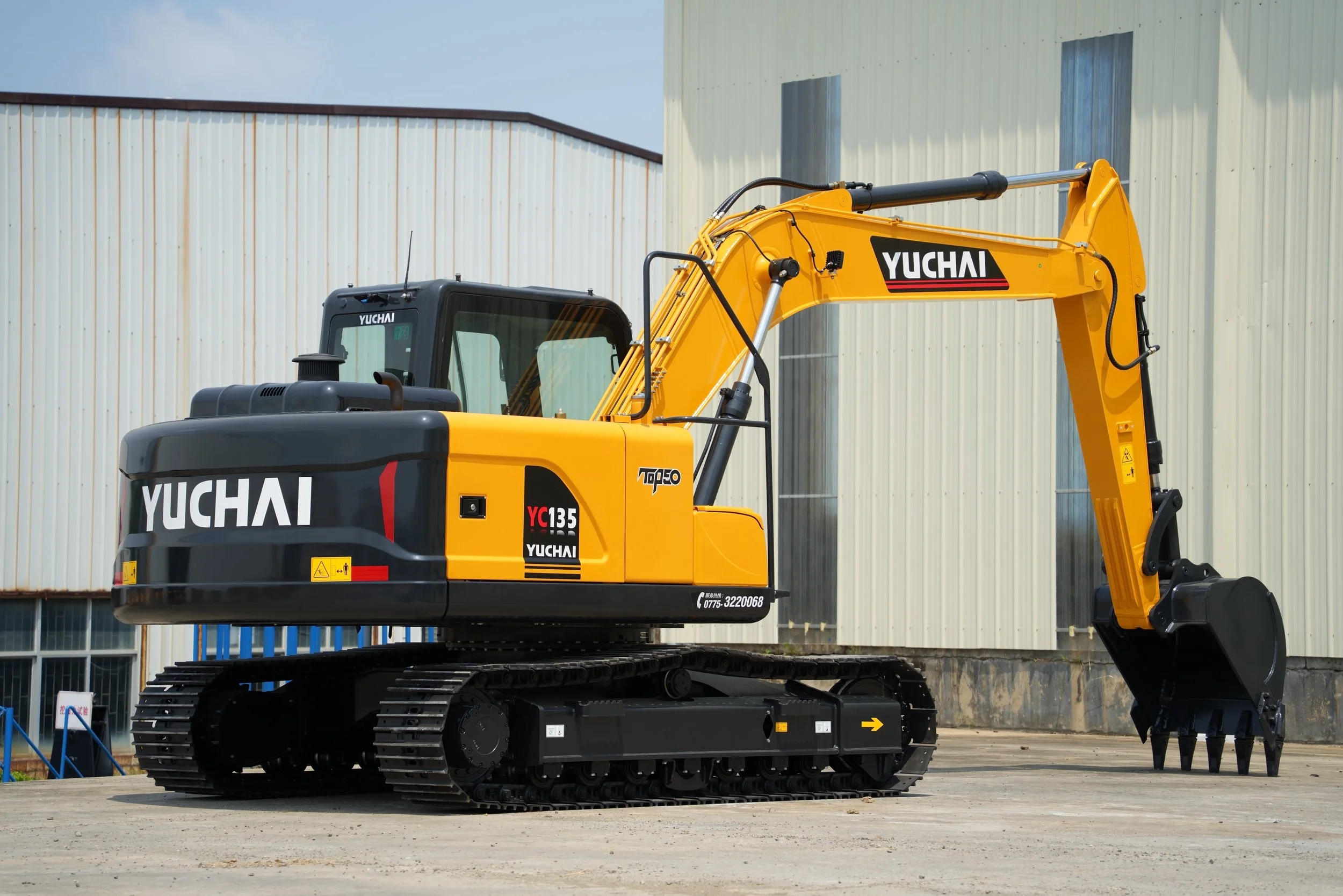 A yellow and black Yuchai excavator parked outdoors in front of a large industrial building with metal siding. The excavator has tracked wheels and a hydraulic arm.