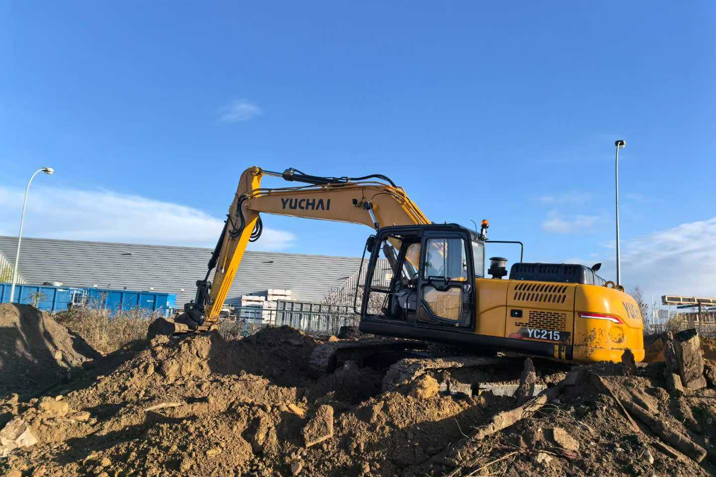 Yellow excavator with 'YUCHAI' written on the arm and 'YC215' on the body, digging a large hole on a construction site, with a clear blue sky above.