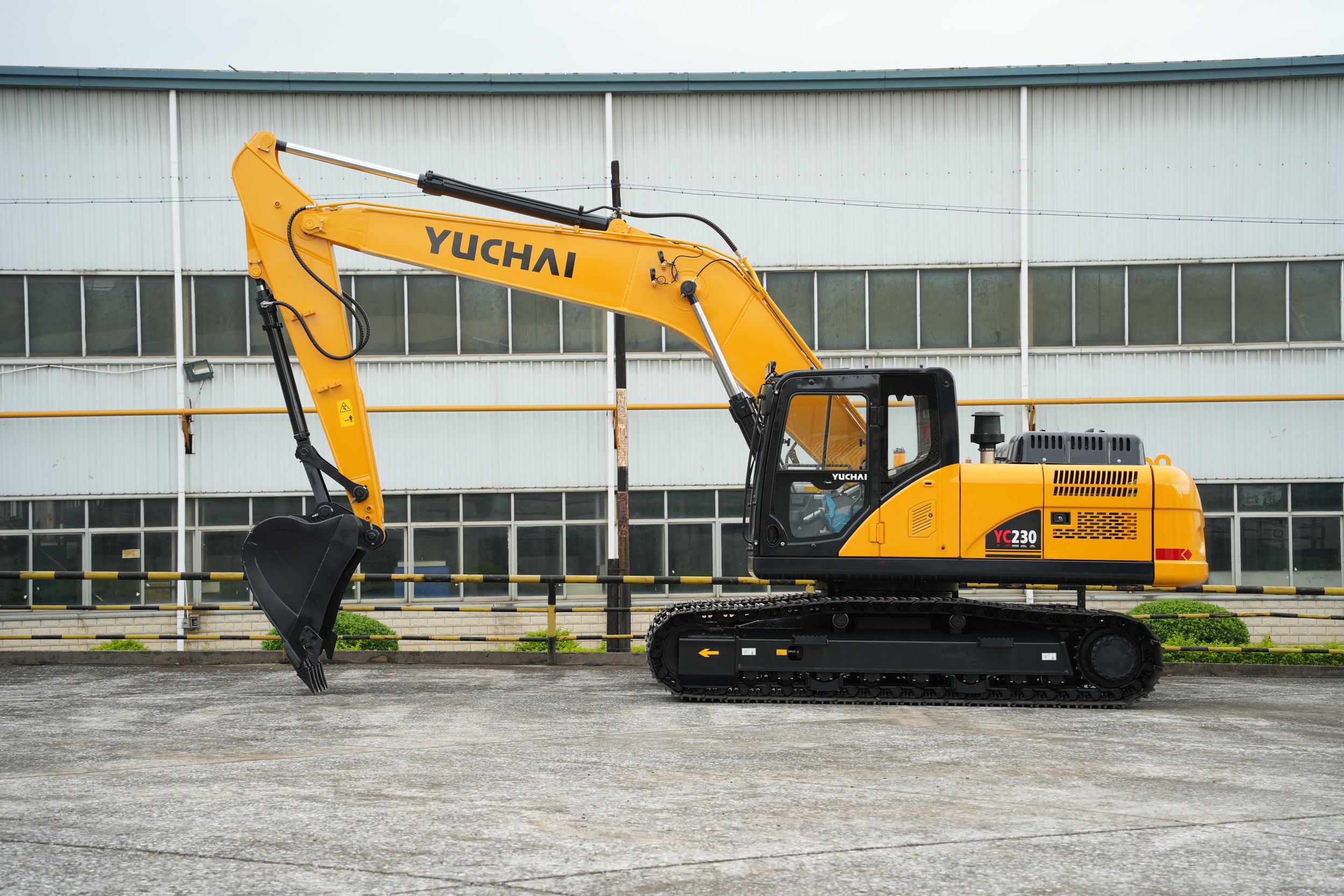 Yellow Yuchai excavator with black tracks parked on a paved surface in front of a modern industrial building with large windows.