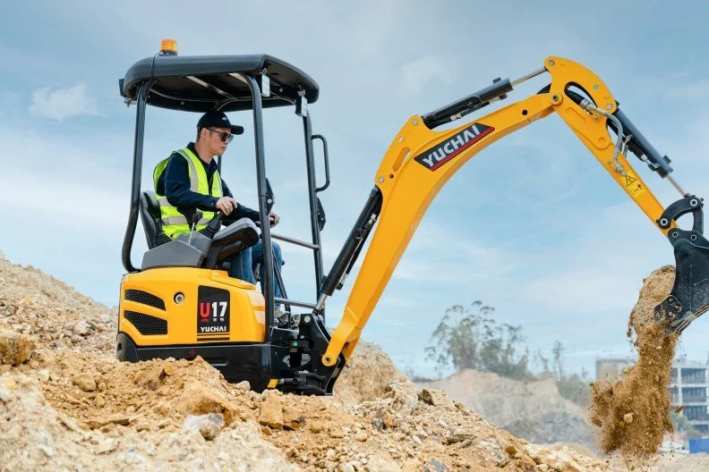 A man operating a yellow Yuchai mini excavator on a construction site with dirt and a blue sky.