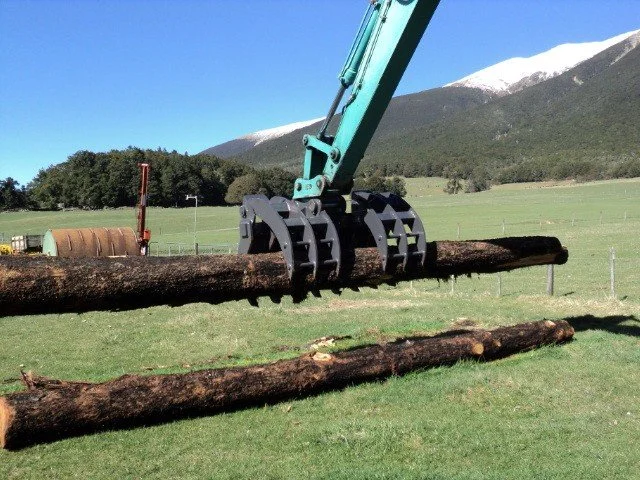 A mechanical clamp attached to a green excavator arm lifting a large log, with another log lying on the ground below in a field with mountains and trees in the background.
