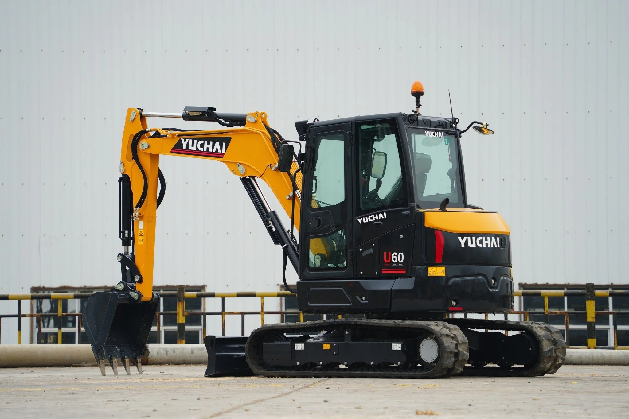 Yellow and black Yuchai mini excavator with a bucket attachment, parked on a paved surface in front of a corrugated metal wall.