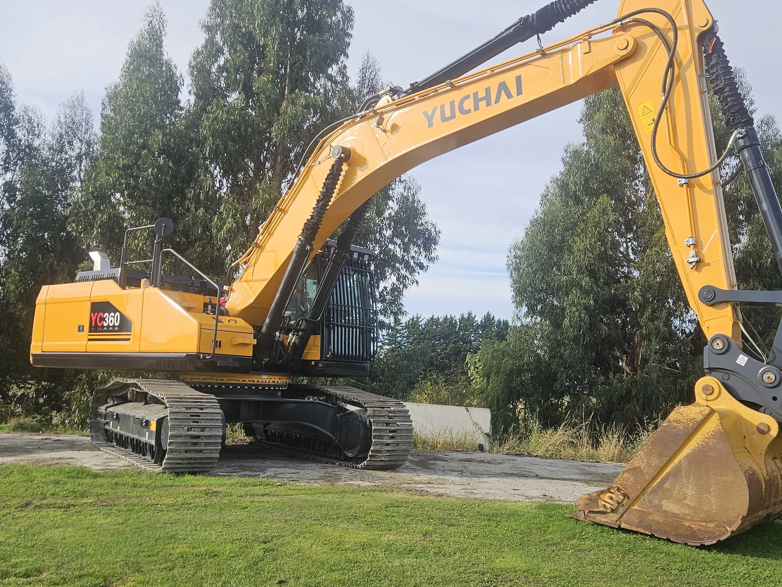 Yellow Yuchai YC360 excavator with a large digging bucket attached, parked on grass and dirt with trees in the background.