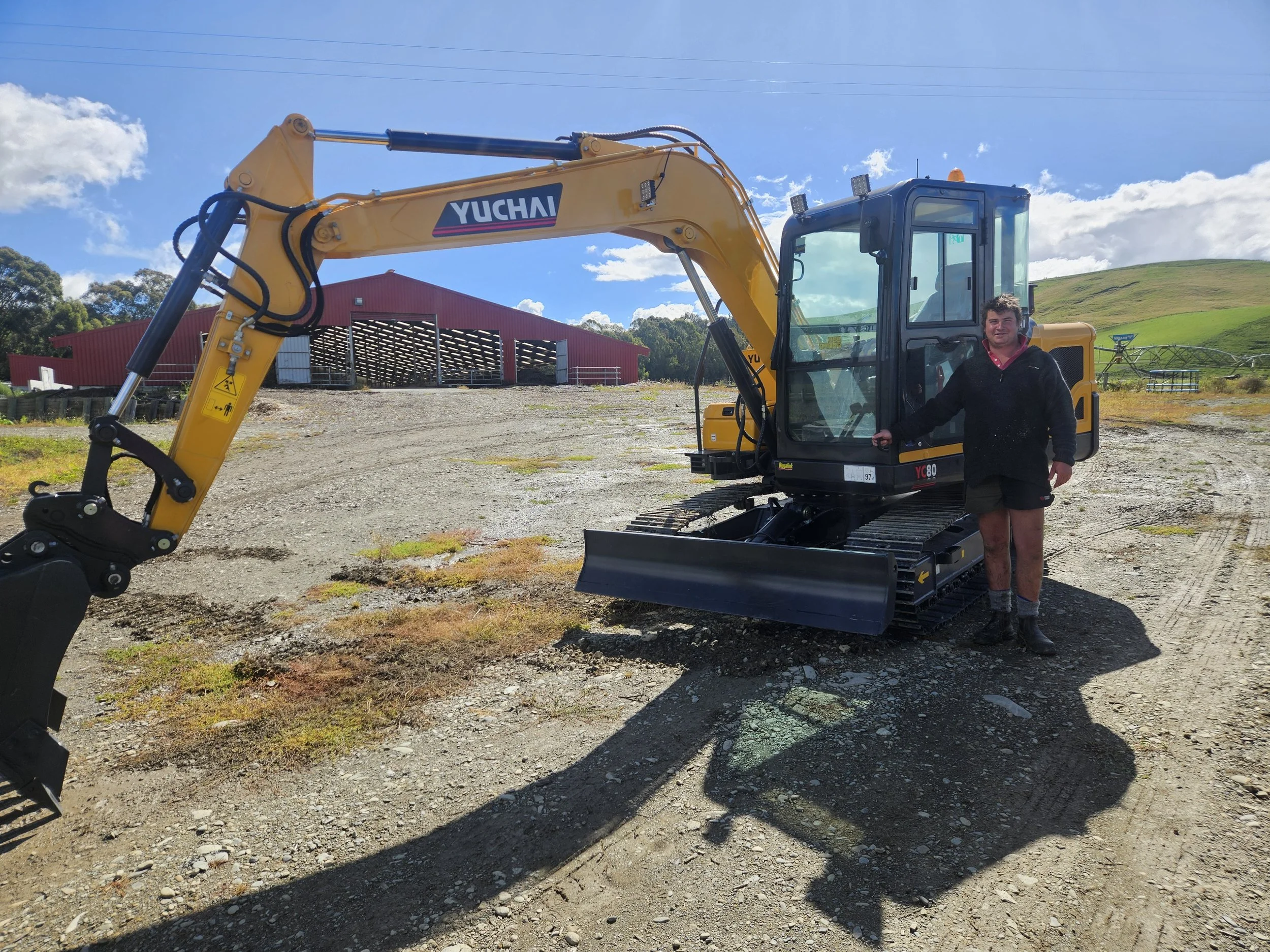 A man standing next to a small yellow excavator on a dirt field with green hills and a barn in the background under a blue sky with scattered clouds.