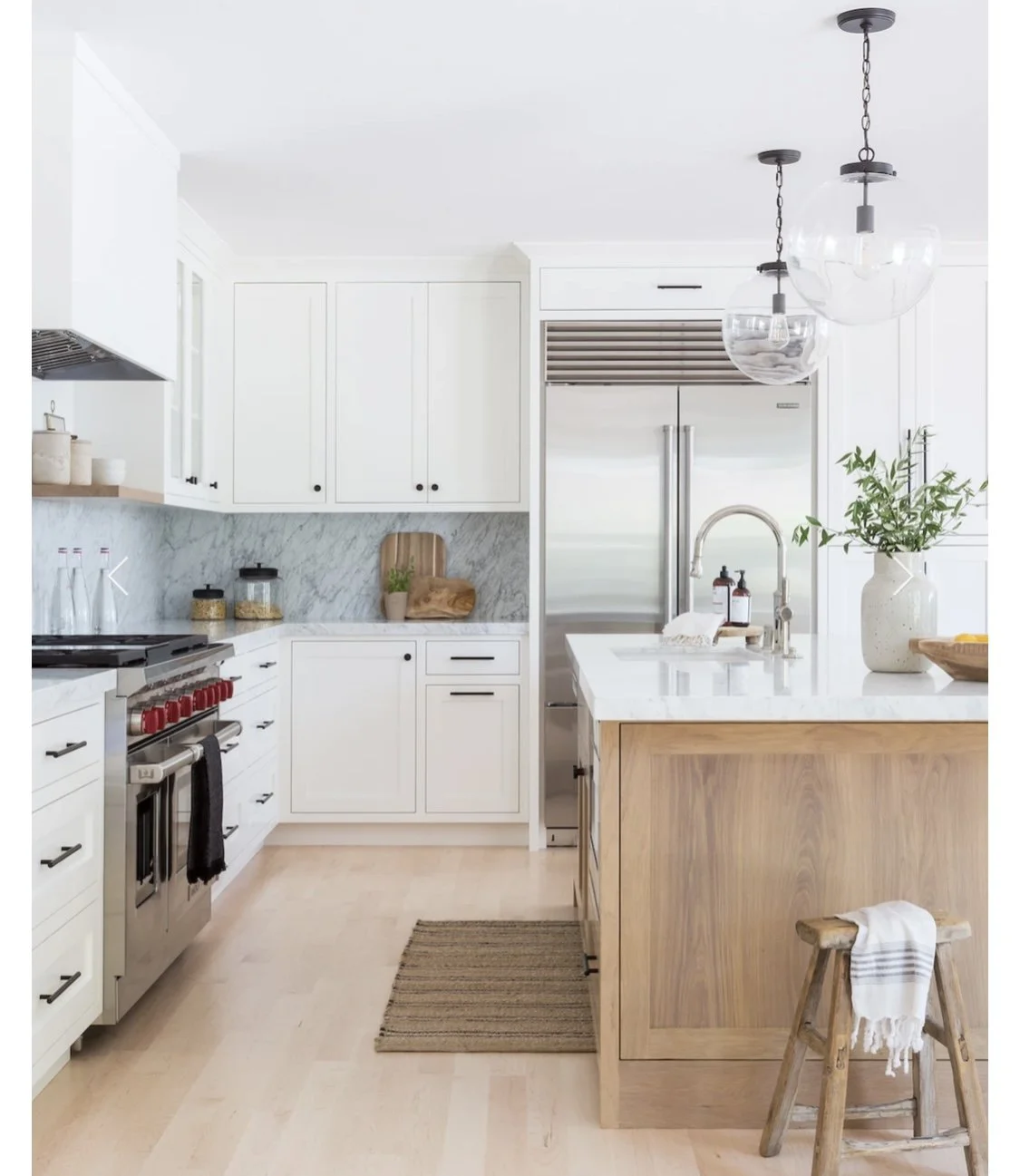 Bright, modern kitchen with white cabinets, marble backsplash, stainless steel refrigerator, wooden kitchen island, and pendant lighting with glass globes. Decor includes potted plant, breadboard, jars, soap dispensers, and a kitchen towel.