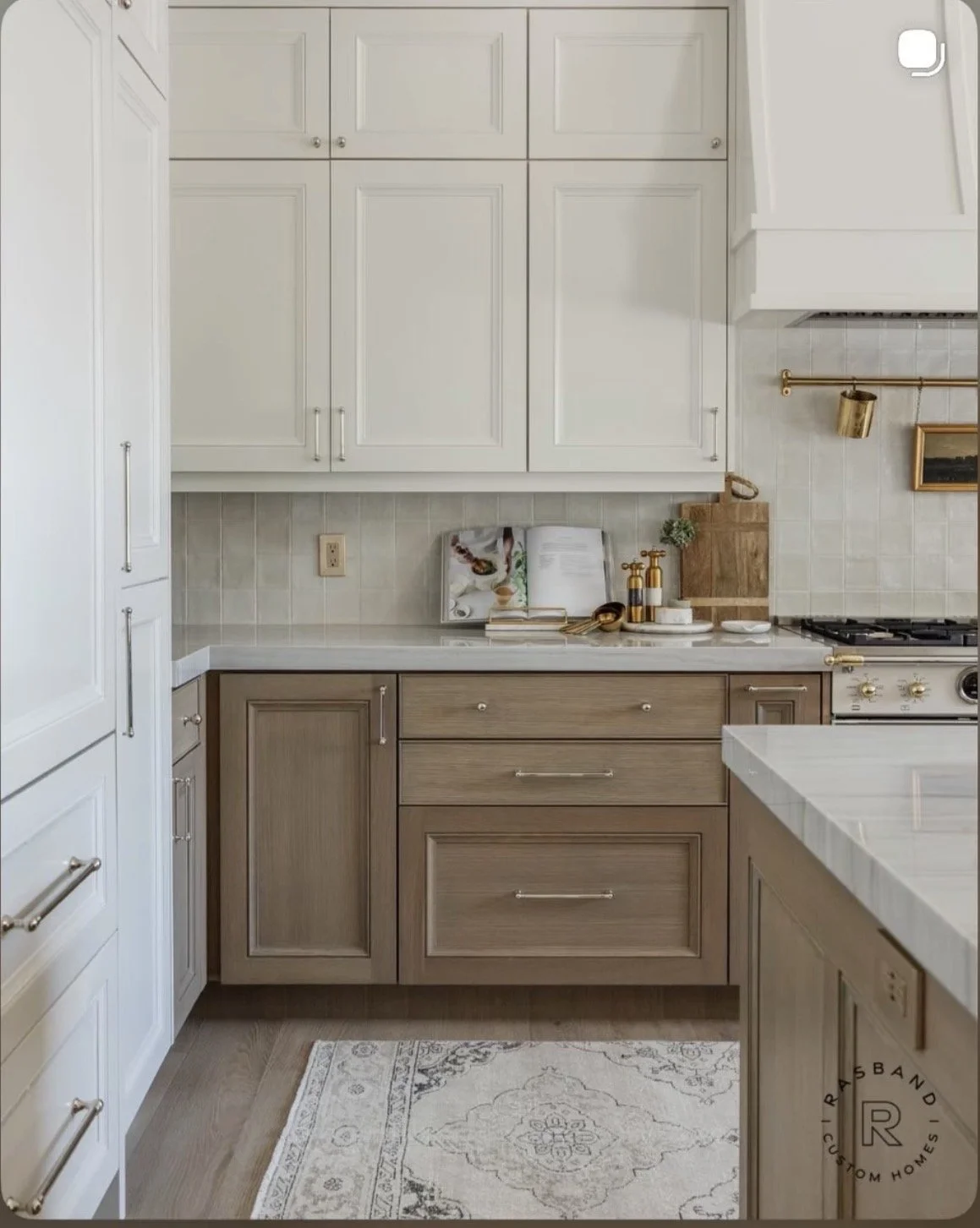 Kitchen with white upper cabinets, light wood lower cabinets, beige tile backsplash, gray countertop, and a white island with gray veining. Decor includes books, gold-toned soap dispensers, chopping board, and a small framed picture.