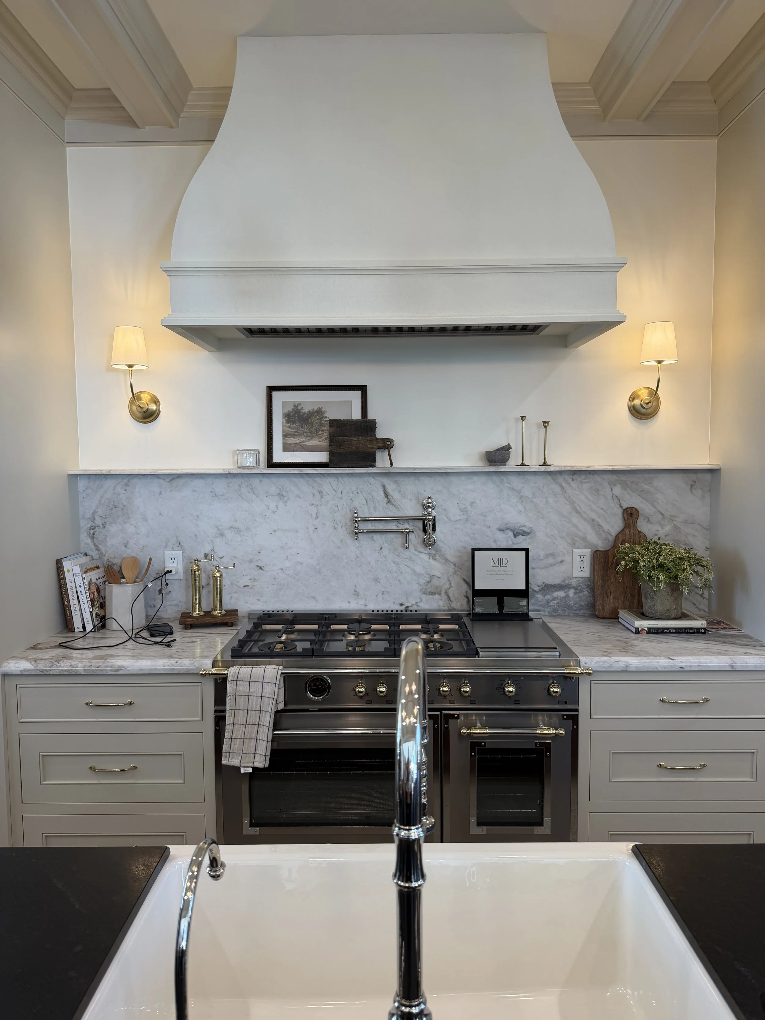 Modern kitchen with white cabinetry, marble countertops, and a gas stove. There are wall-mounted lamps, decorative items, books, and a framed picture above the stove. The image also shows part of a white farmhouse sink and a shiny faucet.