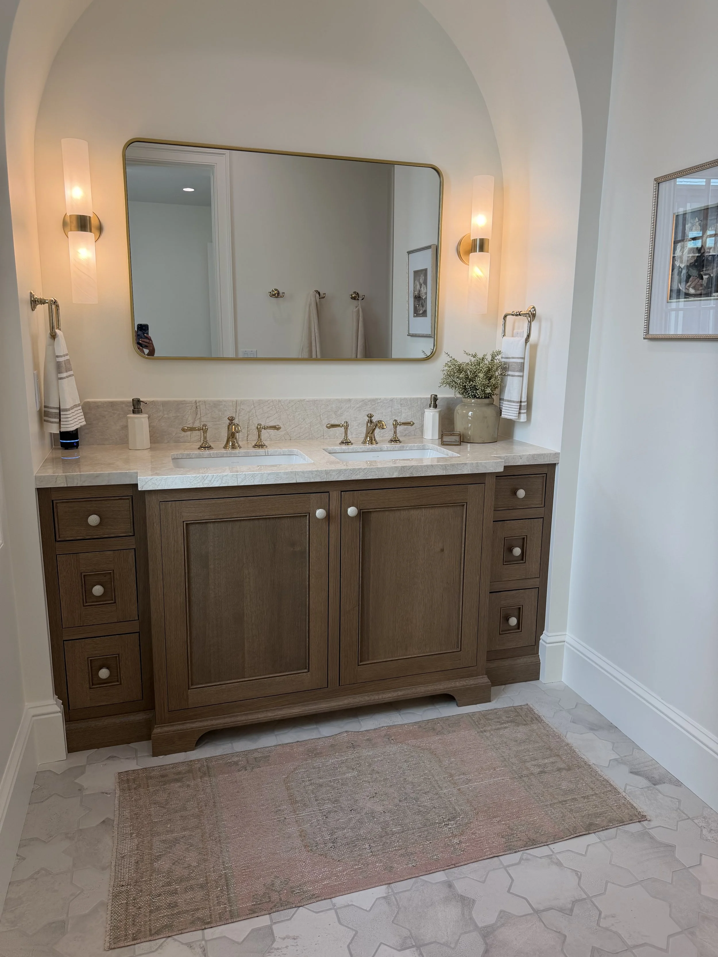 Bathroom with wooden vanity, marble countertop, double sinks, large mirror, wall-mounted lights, towels, a vase with flowers, framed picture, and a pink rug on a tiled floor.
