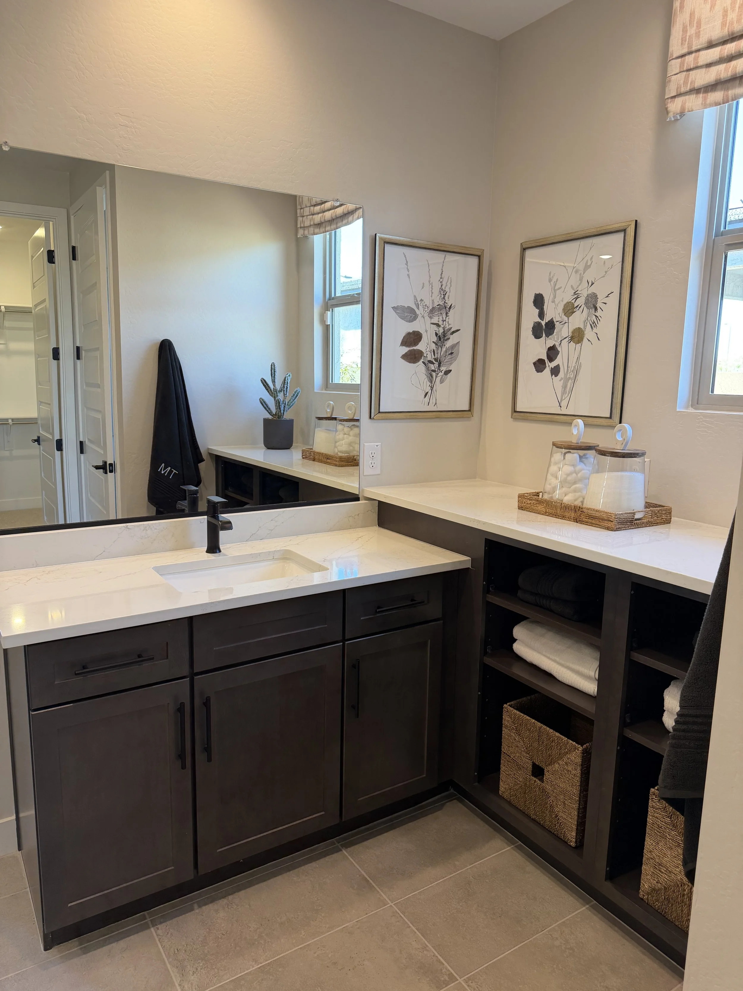 Modern bathroom vanity with dark wood cabinets, white marble countertop, and black faucet. Decor includes framed botanical prints, a small potted cactus, and glass jars with cotton balls. There are open shelves with neatly folded towels and woven sto