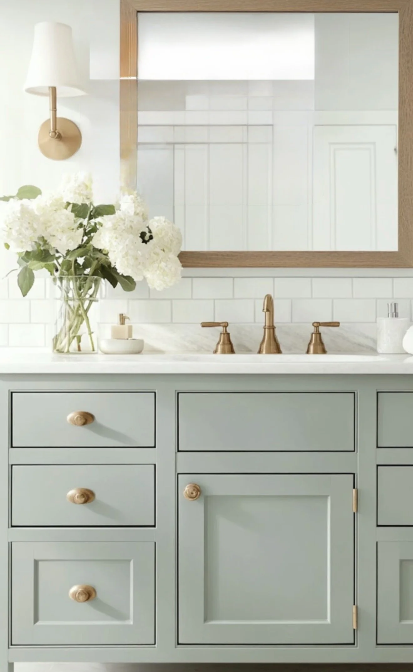 A bathroom vanity with six drawers and two cabinets painted in a soft pastel green color, with round wooden knobs. There is a marble countertop with a gold faucet, a vase of white hydrangeas, a soap dispenser, and a small bowl. A large mirror with a wooden frame hangs above the vanity, reflecting kitchen cabinets, a white wall, and a ceiling light. A wall-mounted light with a white shade is on the left side of the mirror.