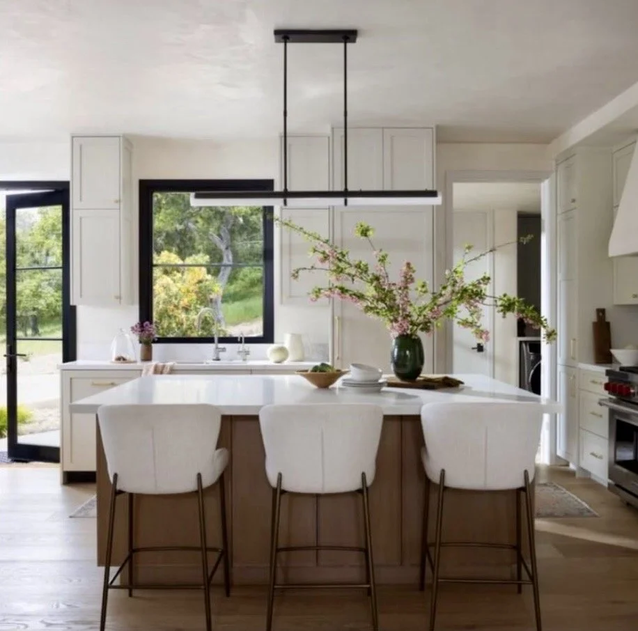 Kitchen with white cabinets, black-framed windows, a large island with bar stools, and a vase of pink flowers on the island.