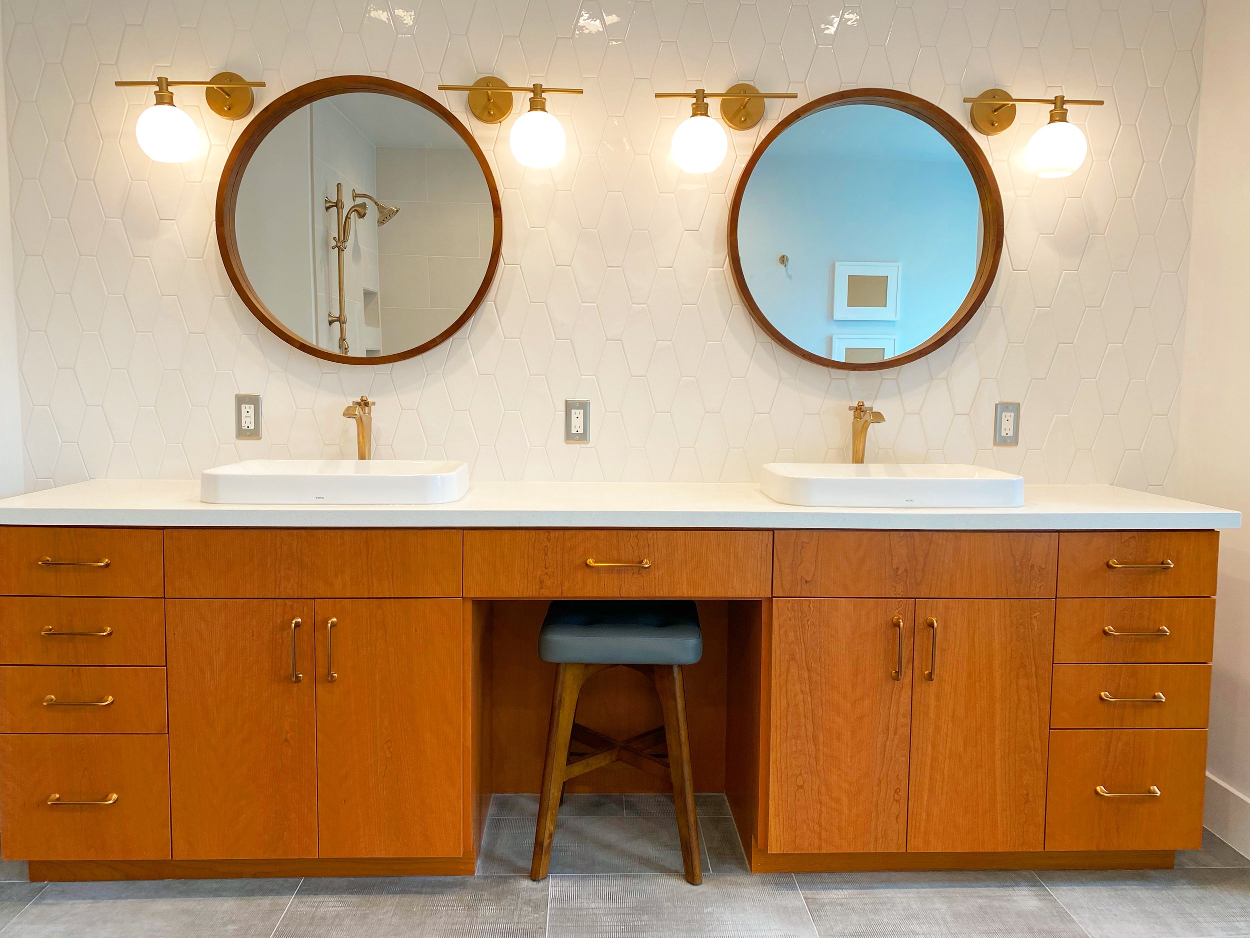 Double bathroom vanity with two round mirrors, white vessels sinks, gold fixtures, wood cabinetry, and a stool in between. Walls are white with a hexagon tile pattern, and there are three light fixtures above each mirror.