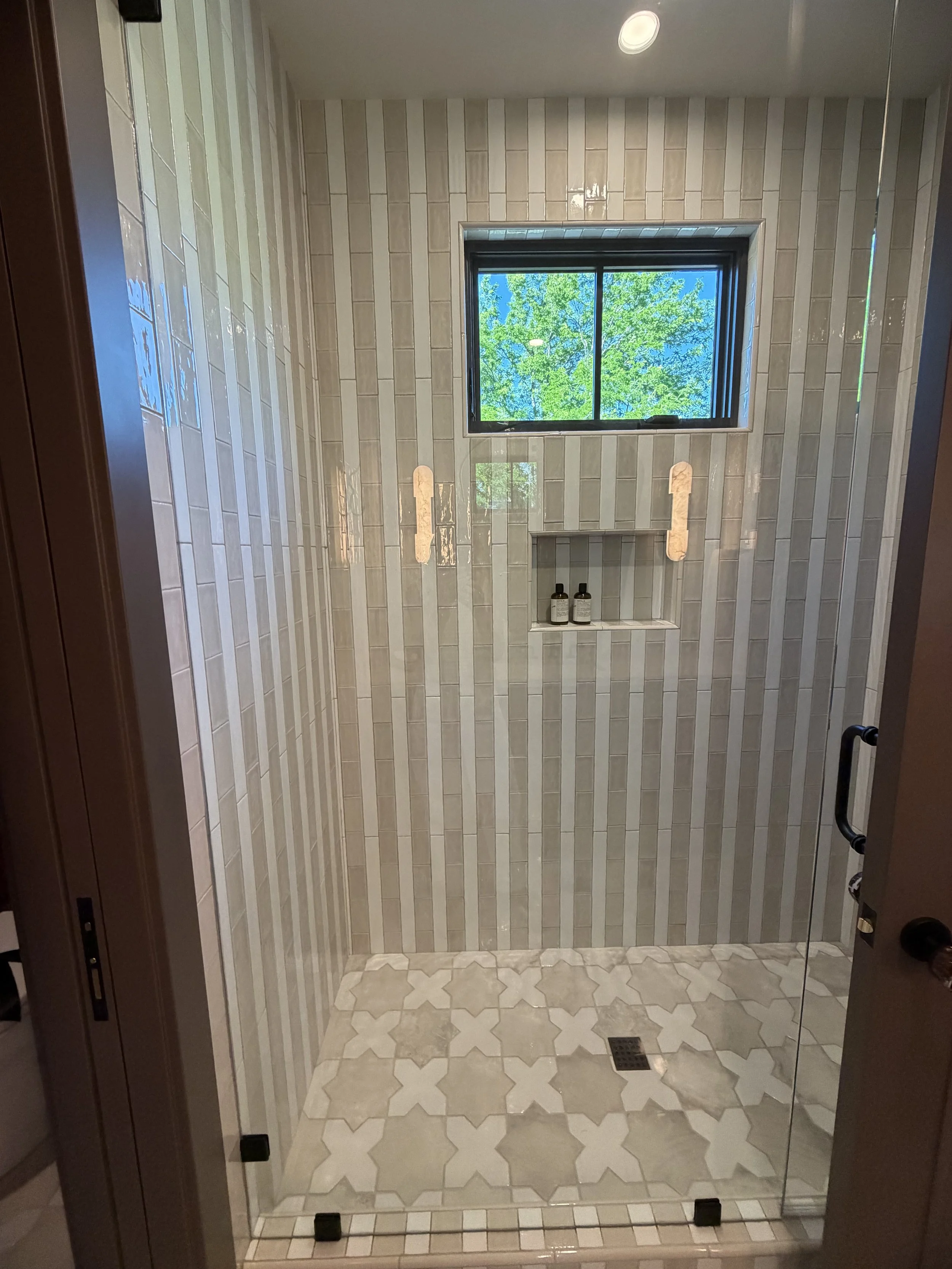 A glass shower enclosure with a tiled interior, a window showing green trees outside, and a tiled floor with a drain.