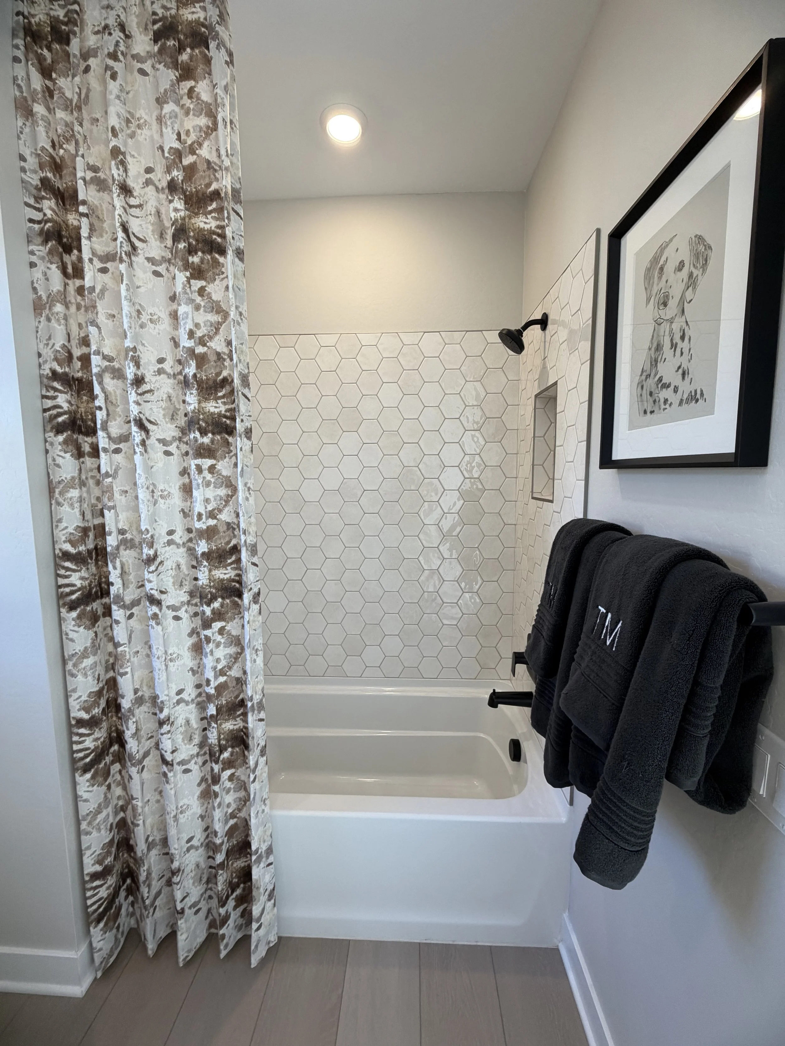 Bathroom shower with white hexagonal tile wall, a bathtub, a black towel rack with black monogrammed towels, decorative shower curtain with beige and brown pattern, framed wall art of a dog, and a recessed light ceiling fixture.