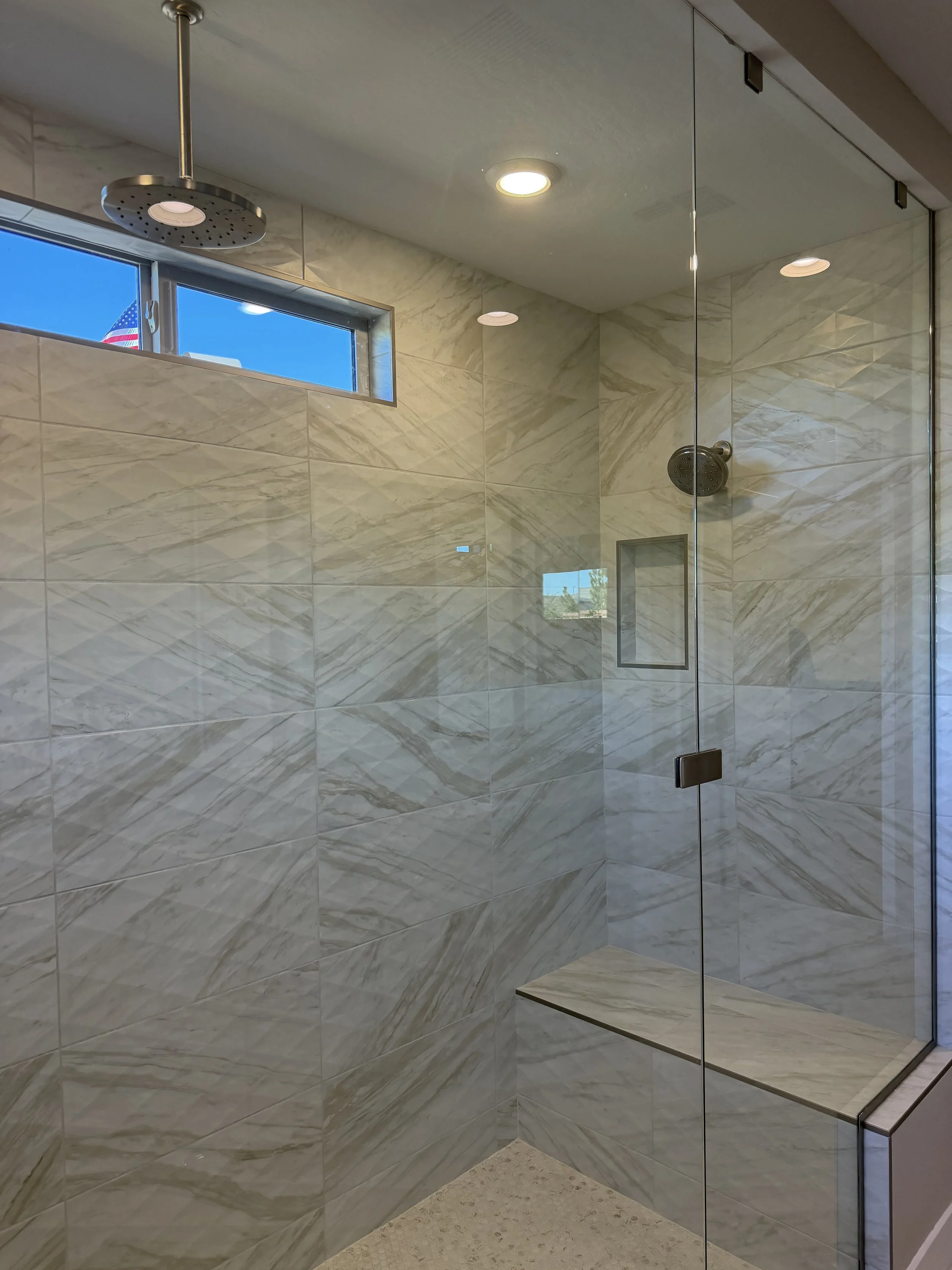 Modern walk-in shower with marble tiles, a small rectangular window near the ceiling, a rainfall showerhead, a separate handheld showerhead, and a built-in bench seat.